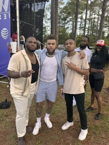 Group of five young adults outdoors in a forested area, posing for a photo at an event, with some making hand gestures and one holding a red cup.