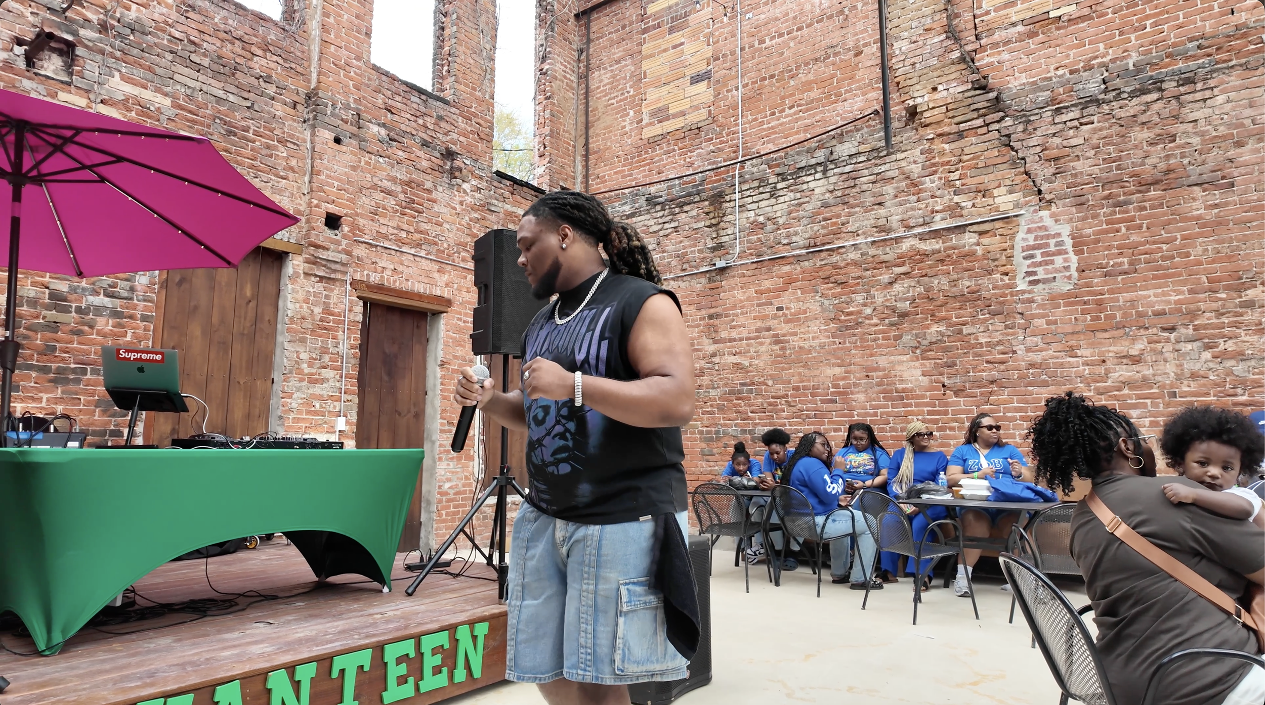 A young man holding a microphone stands on a stage with a DJ setup and a laptop with 'Supreme' logo, in an outdoor venue with exposed brick walls. Behind him, a group of women and children in blue shirts are sitting at tables, some wearing sunglasses