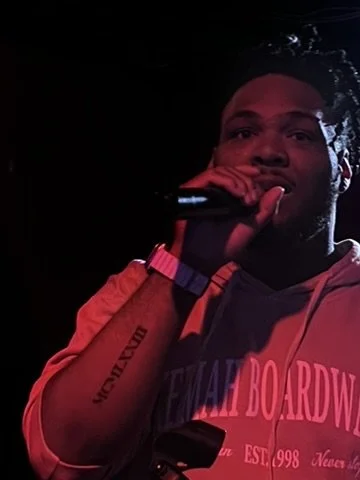 A man holding a microphone in a dark setting, wearing a hoodie with pink text.