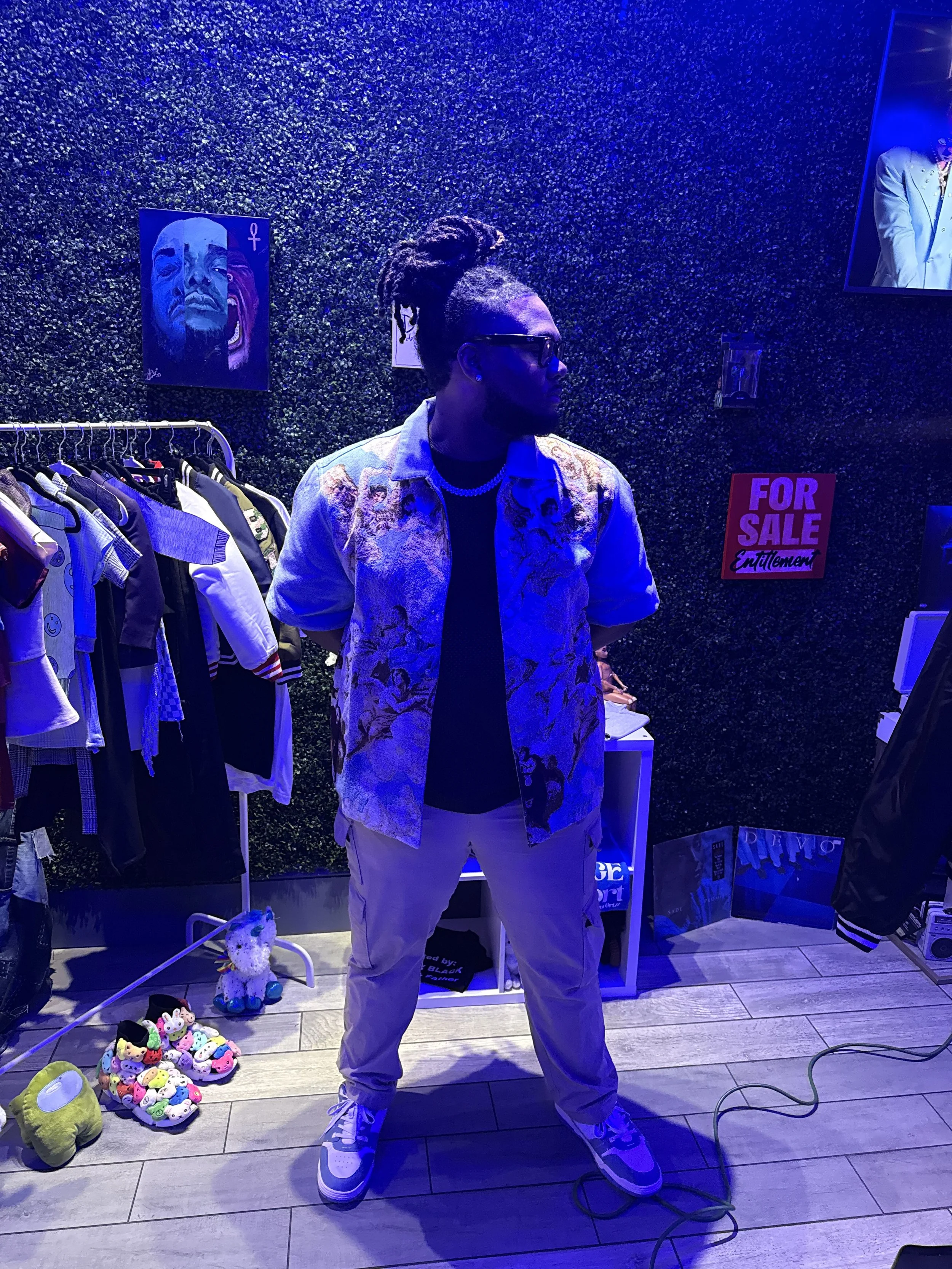 A man with dreadlocks and sunglasses stands inside a store with a textured dark green wall, clothing rack, colorful toys, and a 'For Sale' sign.