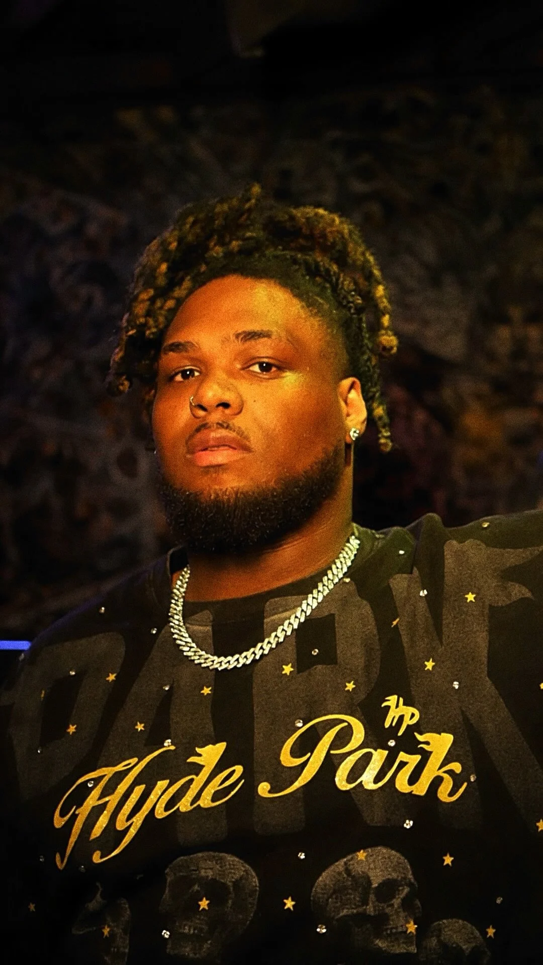 A young man with dreadlocks styled in an updo, wearing a black shirt with gold lettering and skull motifs, silver chains, and earrings, posing against a dark, textured background.