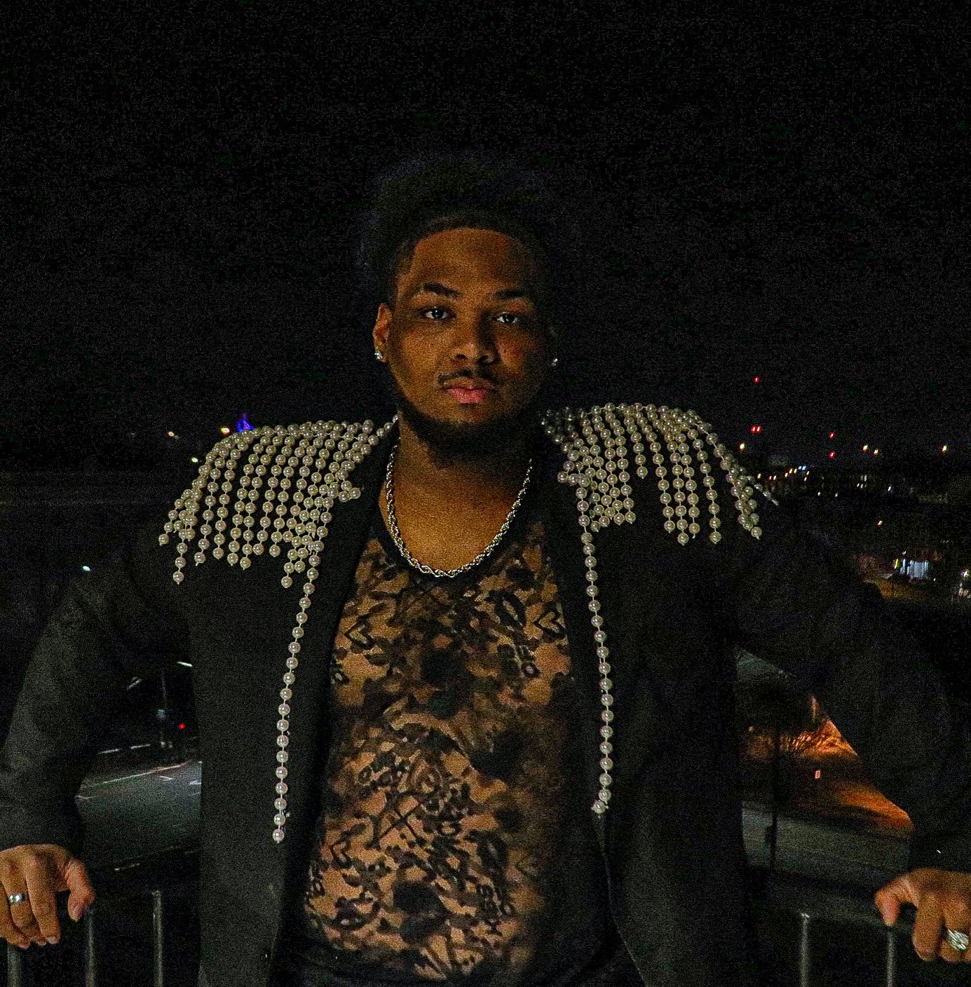 A man wearing a black jacket with pearl embellishments on the shoulders and a leopard print lace shirt stands at night with a city skyline lit up in the background.