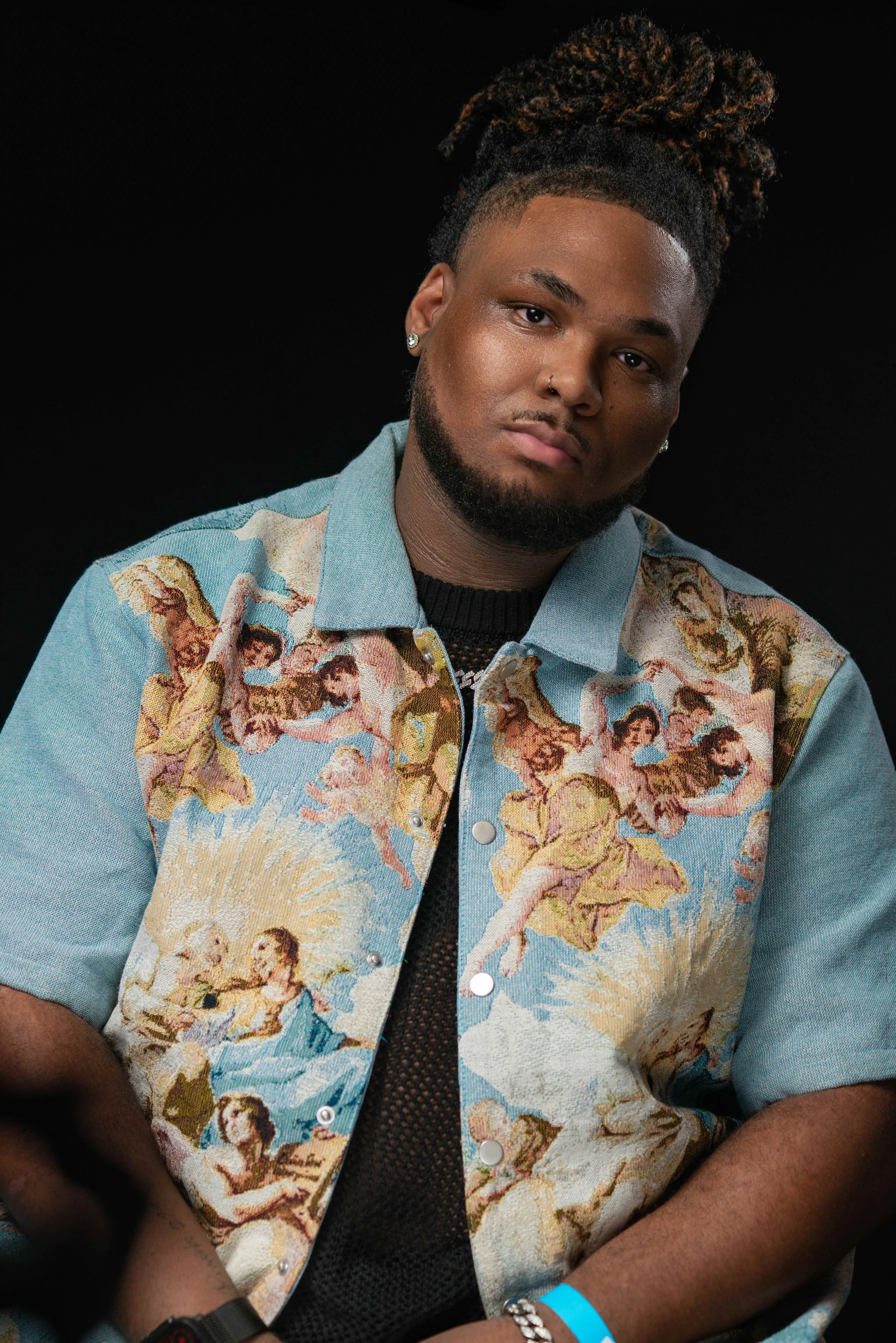 Portrait of a young man with dreadlocks in a high bun, wearing a colorful jacket with biblical-themed artwork, earrings, and a black mesh shirt underneath, against a black background.