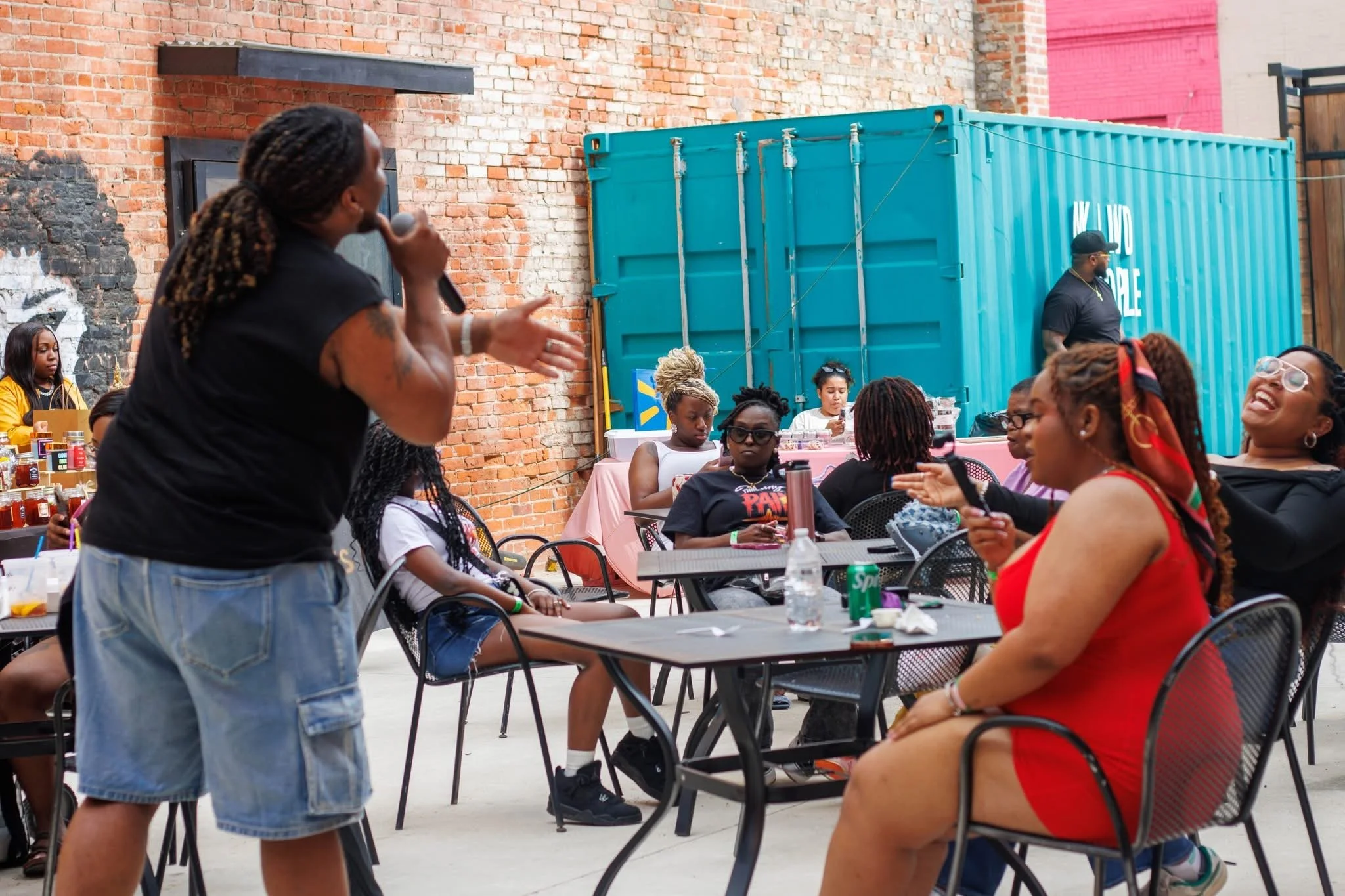 A woman with long braids in a black T-shirt and ripped jeans holds a microphone and gestures toward a group of seated women at an outdoor event with a brick wall and a large blue shipping container in the background.