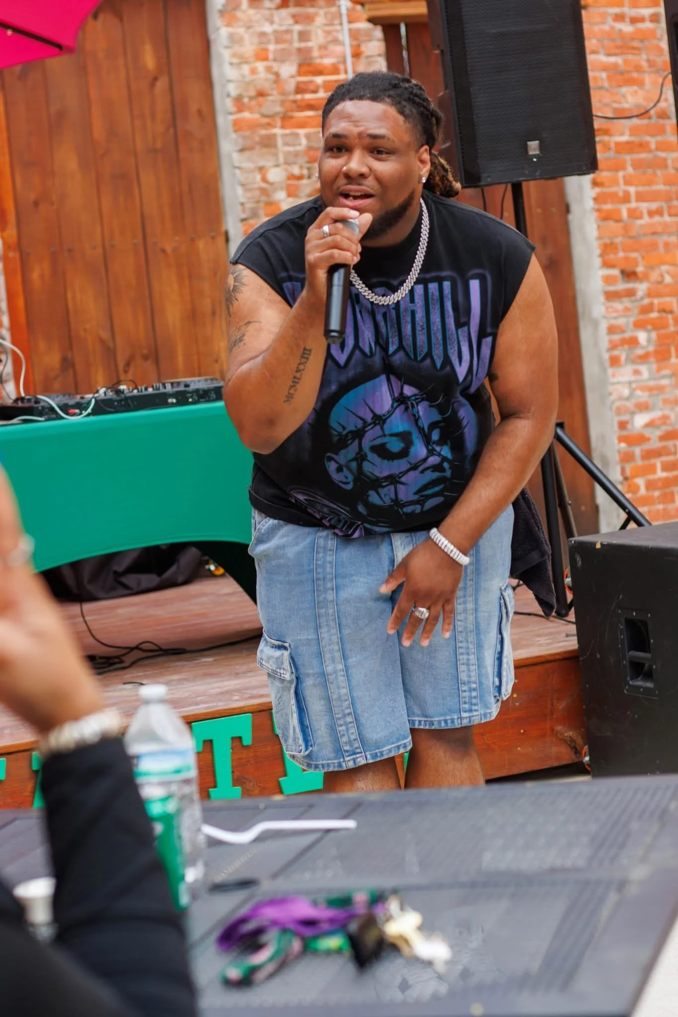 A man with dreadlocks, wearing a black sleeveless graphic T-shirt, denim shorts, and jewelry, holding a microphone and performing on stage at an outdoor event.