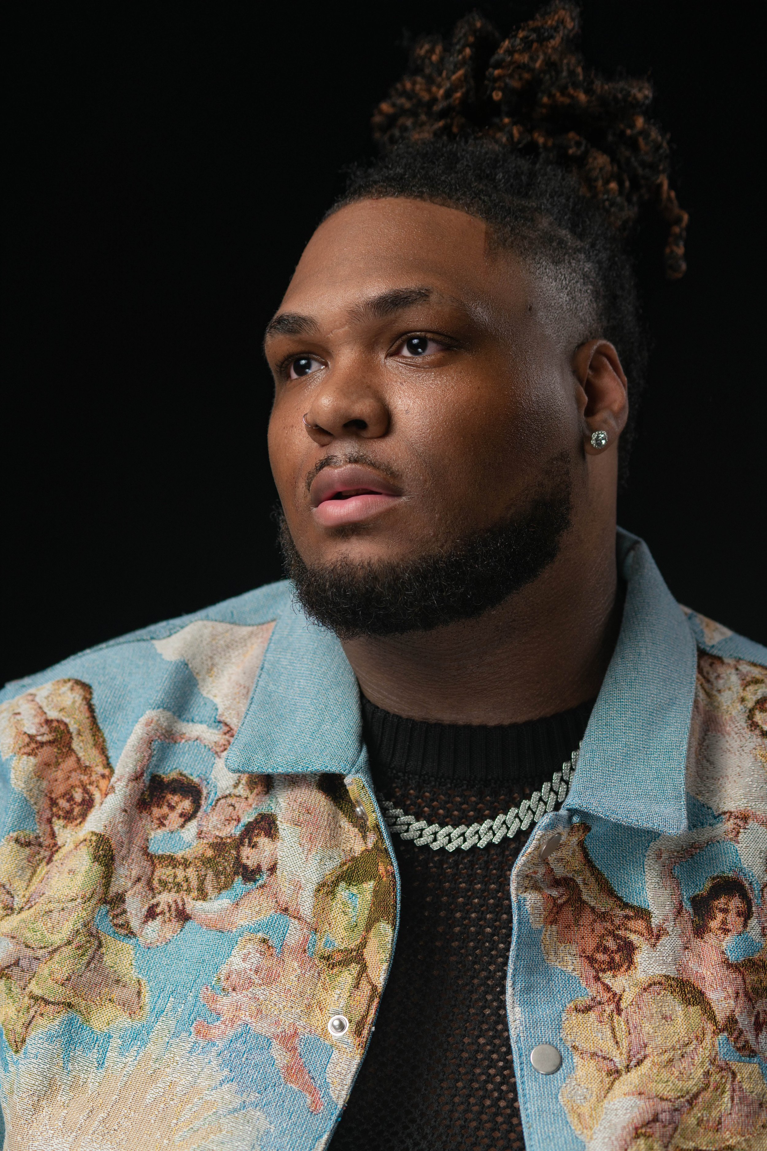 Portrait of a young man with a beard, earrings, and styled hair, wearing a colorful patterned jacket and a black mesh top, against a dark background.