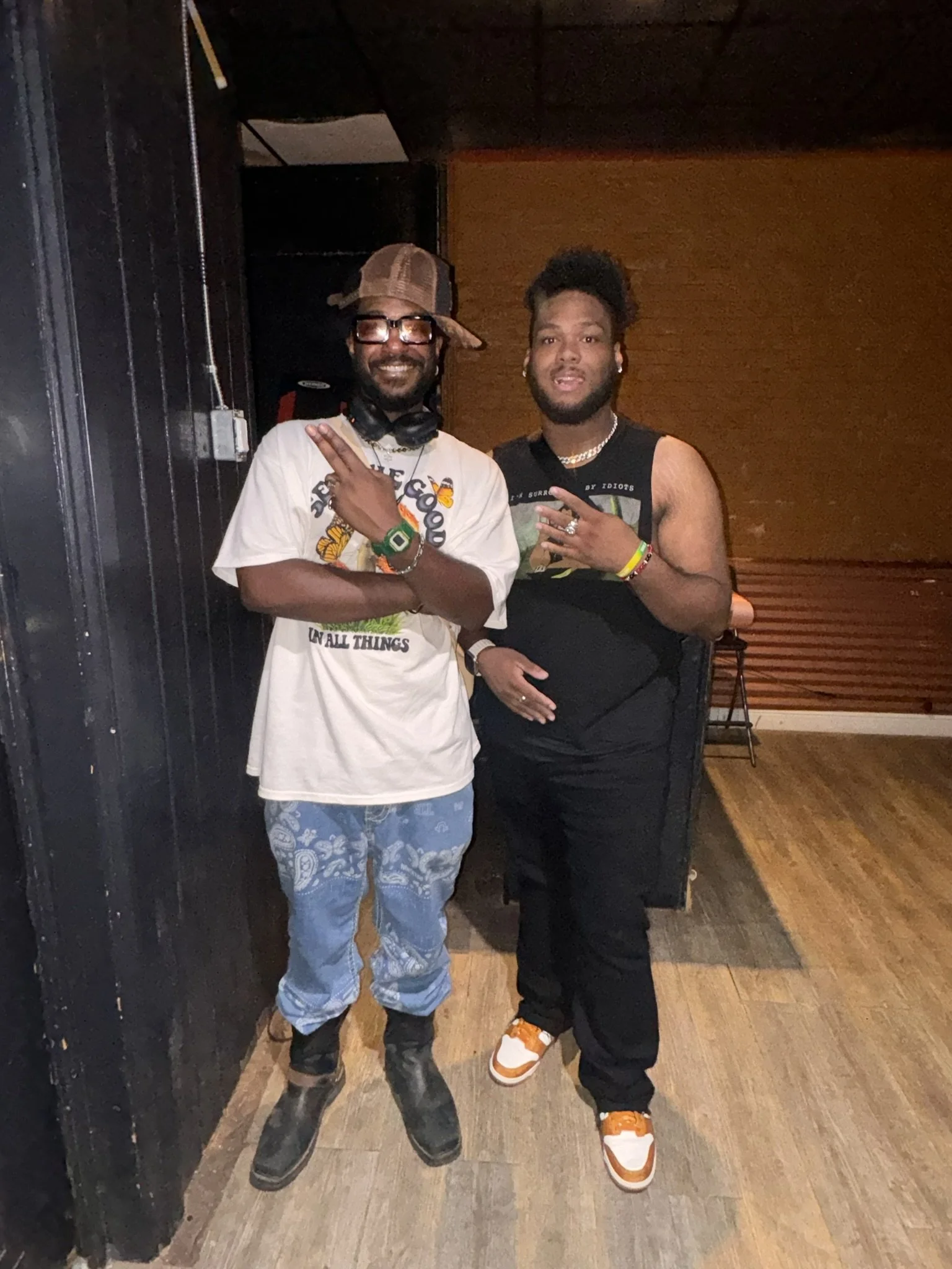 Two men standing indoors, smiling and making hand gestures. One wears a graphic T-shirt, blue patterned pants, glasses, a hat, and boots. The other wears a sleeveless shirt, black pants, and sneakers, with jewelry and an afro hairstyle.