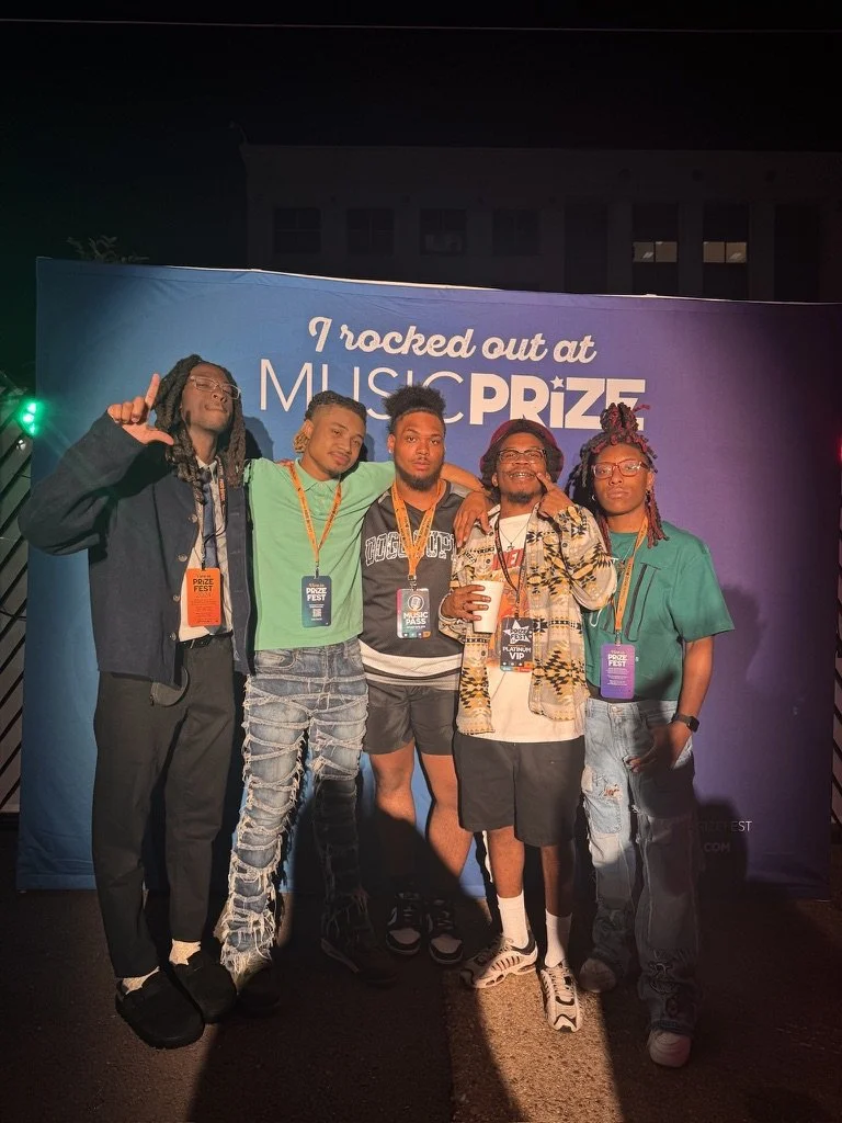 Five young men standing together at night in front of a blue backdrop that says "I rocked out at MUSIC PRIZE." They are wearing casual clothing and have event passes around their necks, smiling for the photo.
