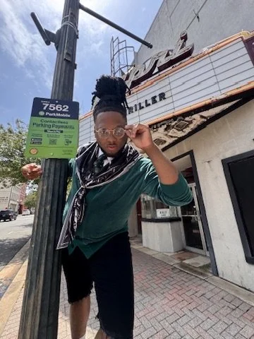 Person standing on sidewalk adjusting sunglasses near a parking meter and a streetlamp with a building and marquee in the background.