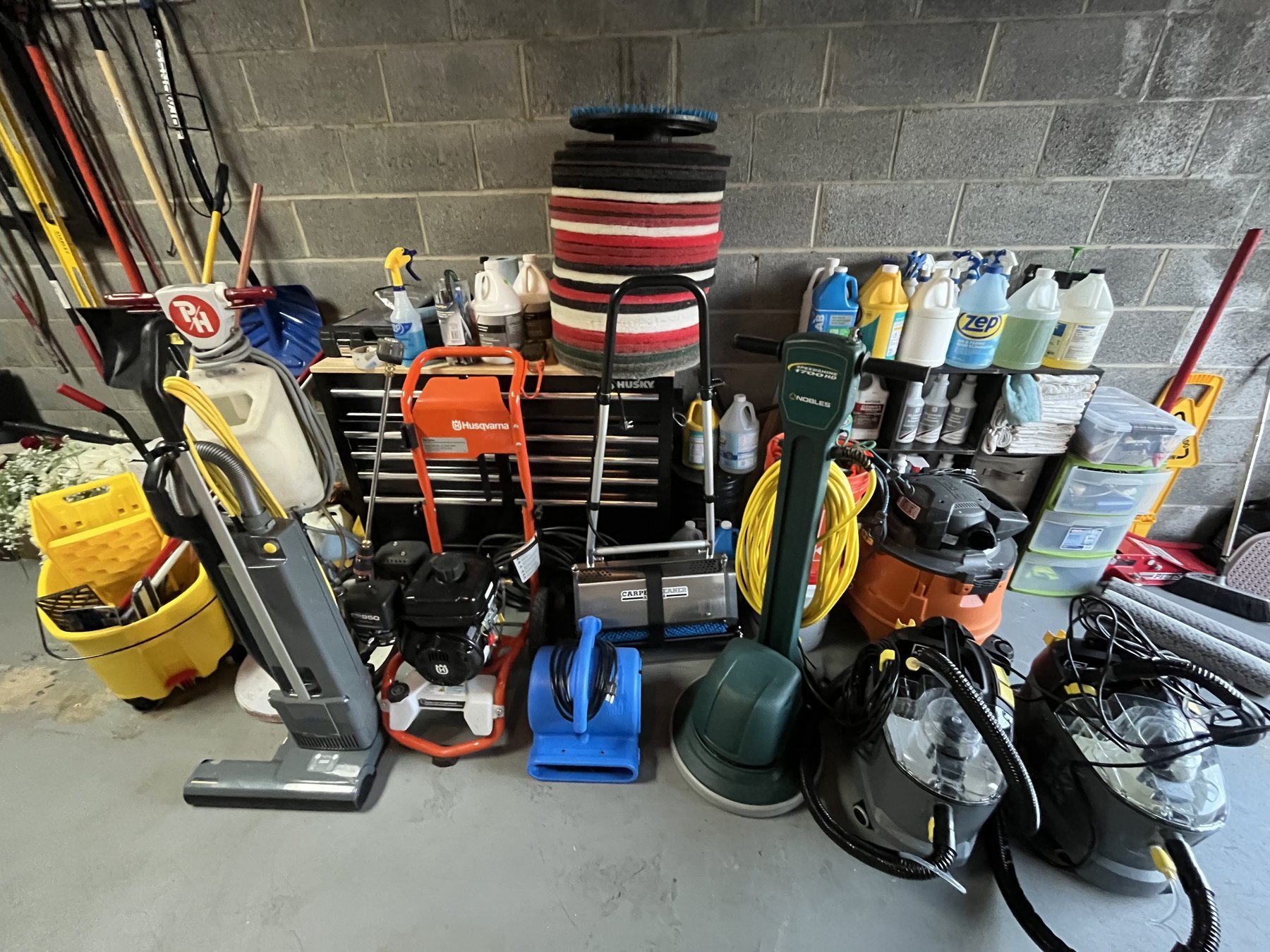 Lumina Surfaces equipment lined up in the shop: a floor buffer, pressure washer, carpet extractors, air movers, stacked floor pads and shelves of cleaning solution.