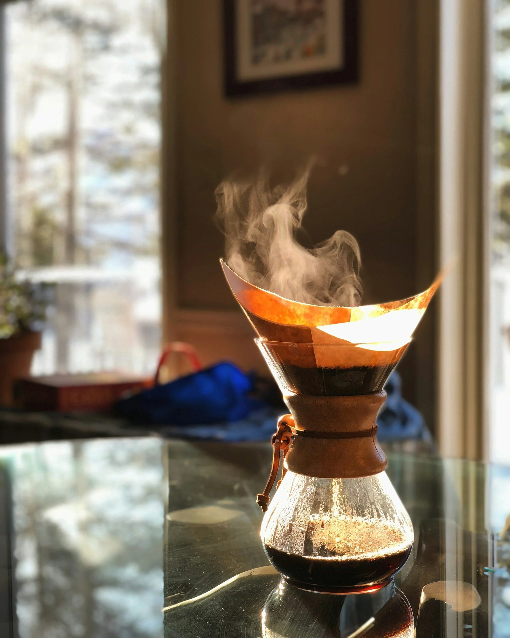 A pour-over coffee maker with steam rising from the brewing coffee, sitting on a glass table in a well-lit room.