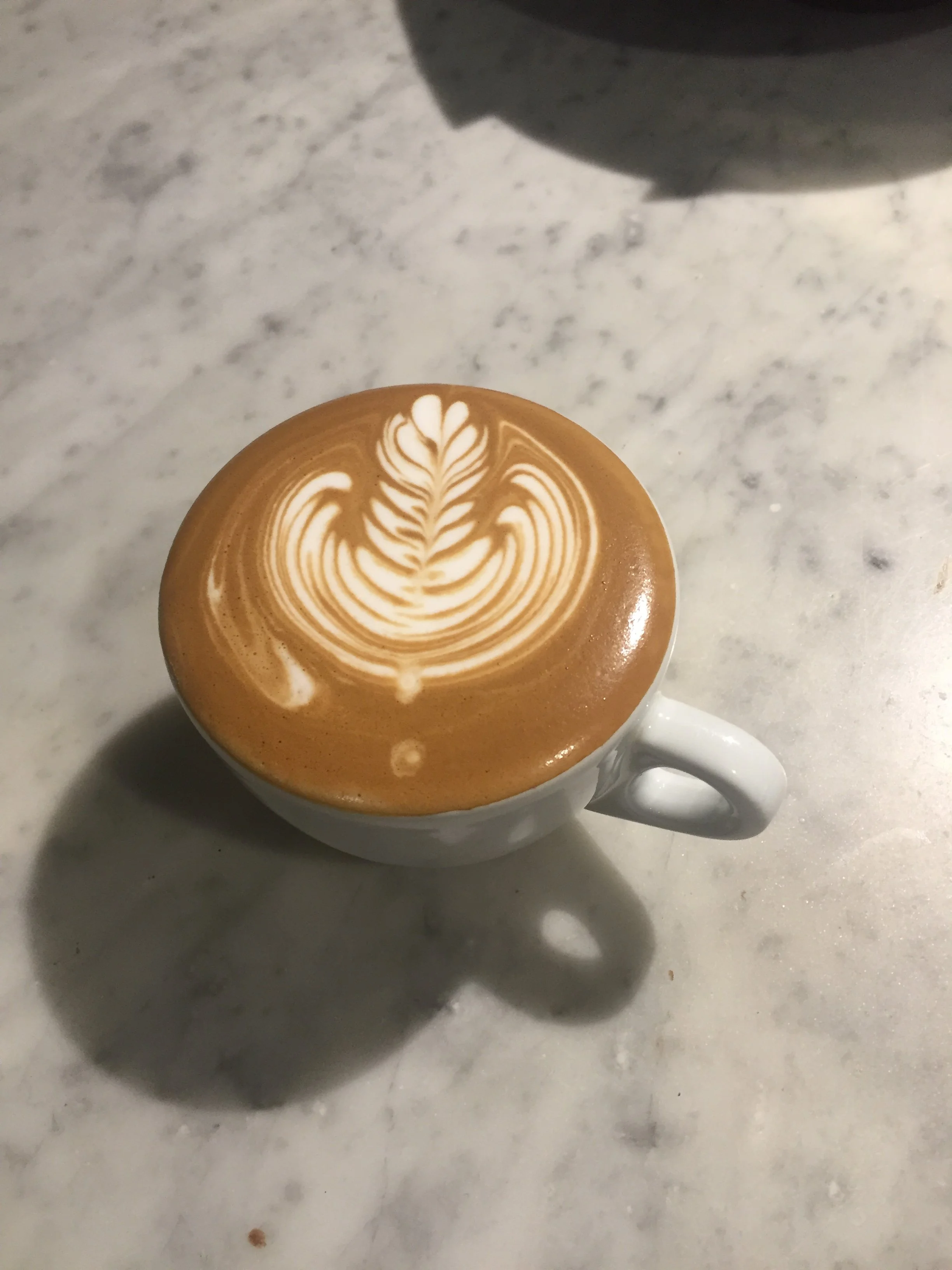 A cup of coffee with latte art in a white mug on a marble table, showing a symmetrical leaf pattern in the foam.