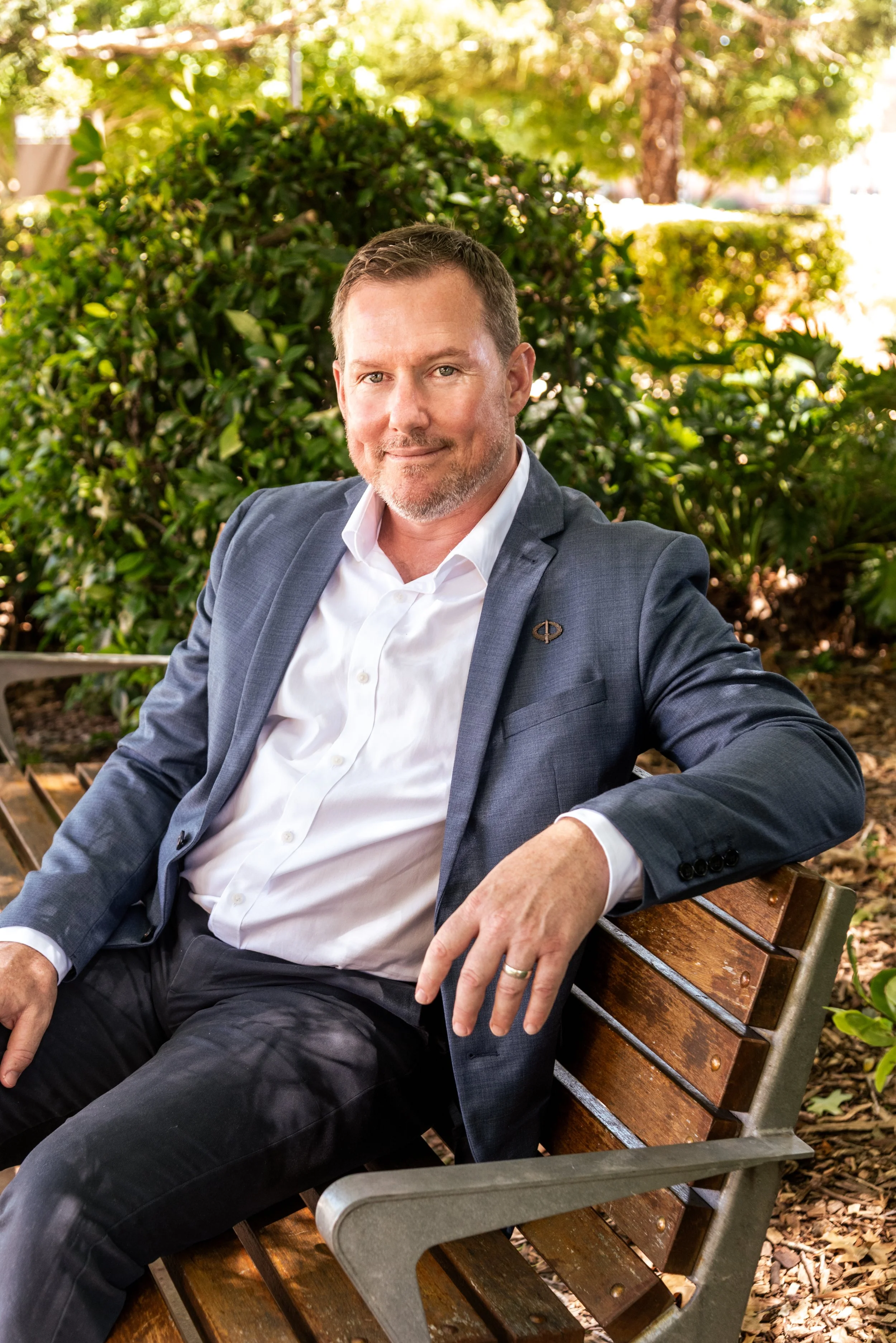 Male, David, wearing a navy blue blazer sitting on a park bench in a relaxed stance with a soft smile.