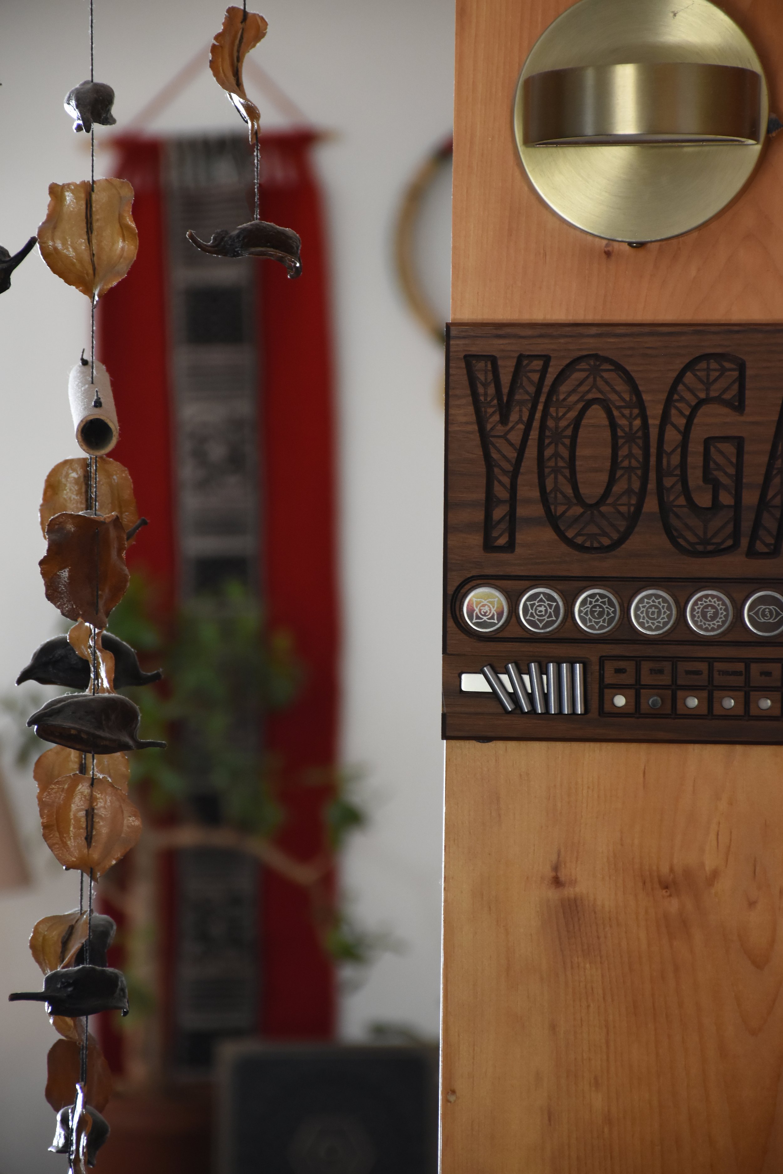 A wooden yoga timer with a metal scale on top, a carved wooden panel with 'YOGA' engraved on it, and six small tokens with symbols, alongside a hanging string of dried fruit and herbs in the background.