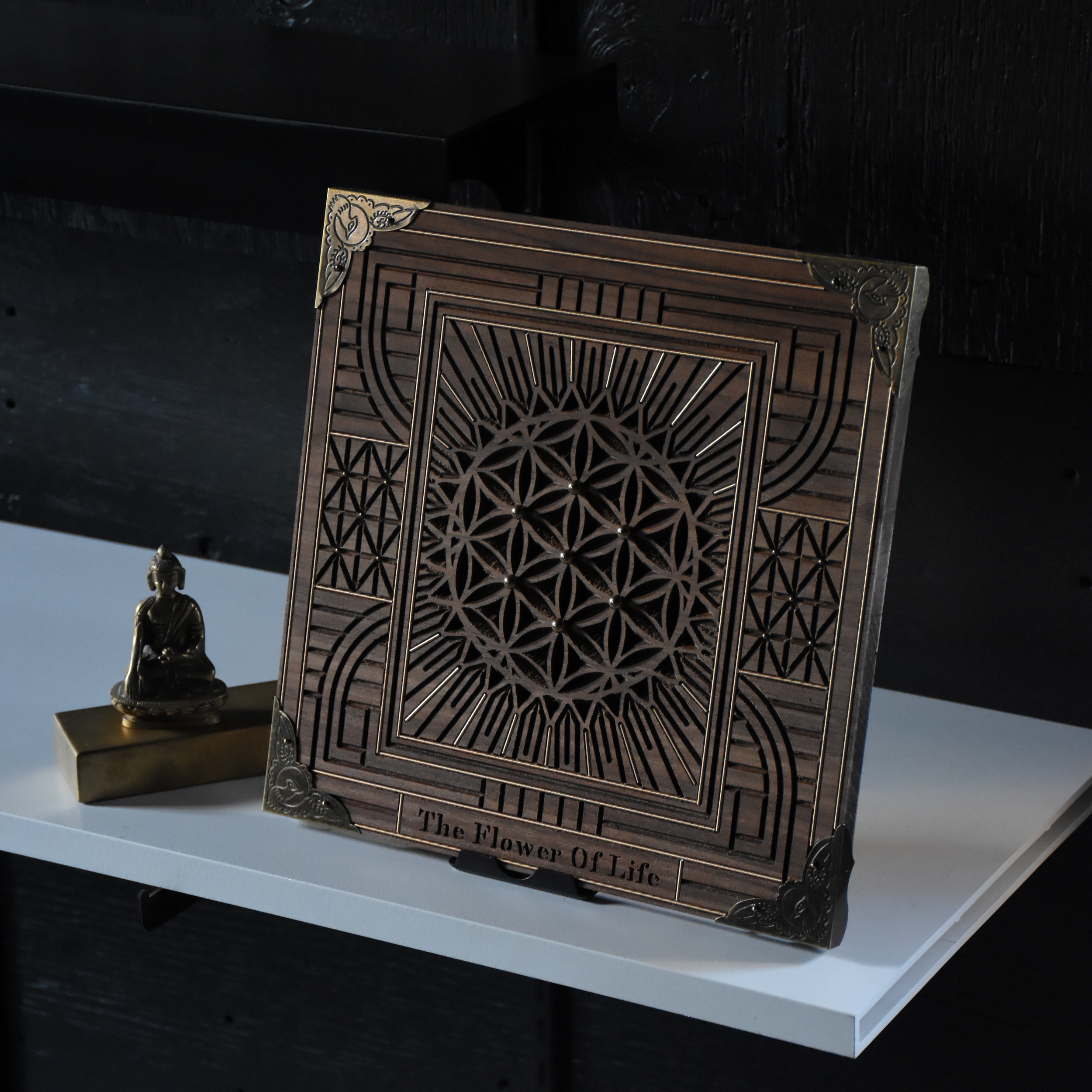 A decorative wooden box with intricate geometric cutouts and a floral design on the corners, labeled 'The Flower Of Life.' Next to it is a small brass Buddha figurine seated in a meditative pose. Both items rest on a white surface against a dark back