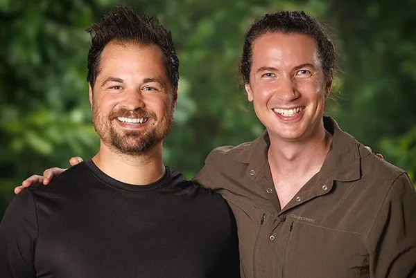 Two men smiling, standing close together outdoors with a green, blurred natural background.