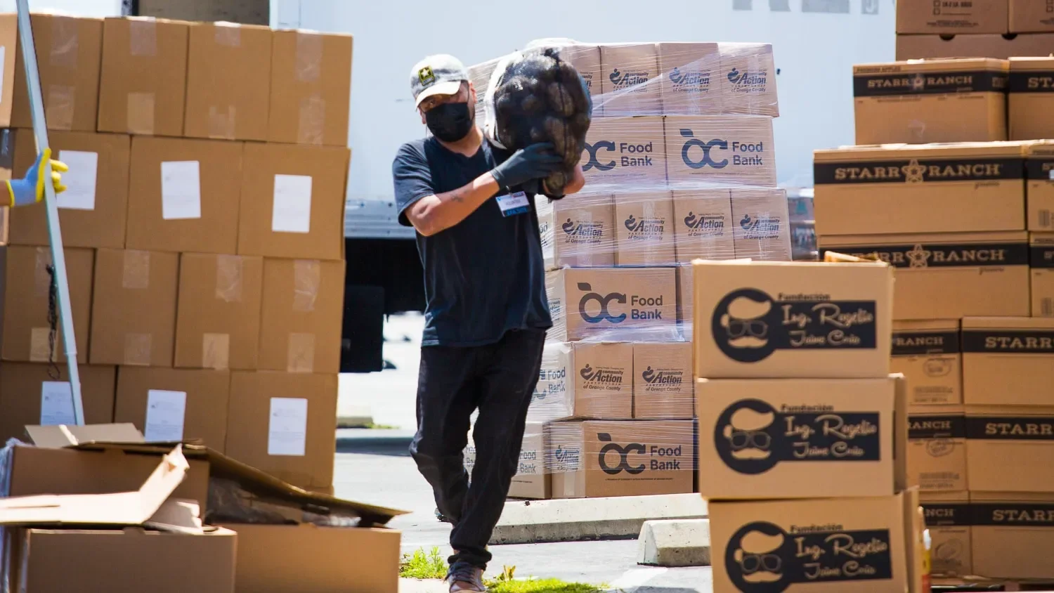 A man wearing a black face mask, black gloves, and a baseball cap loads a large bag into a truck surrounded by stacks of boxes labeled with Food Bank logos and other packaging.