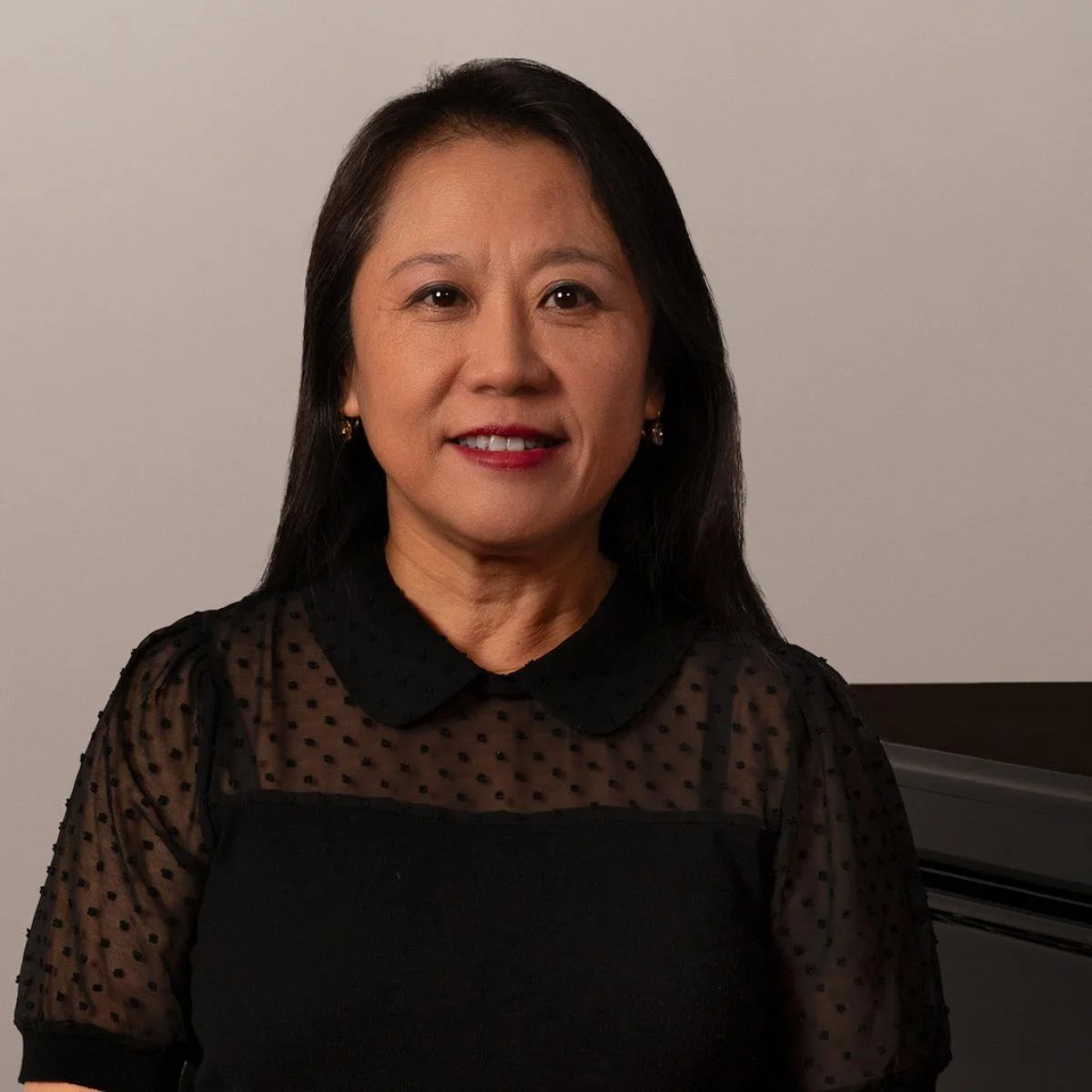 A woman with long black hair wearing a black blouse with sheer polka dot sleeves, sitting in front of a plain white background.