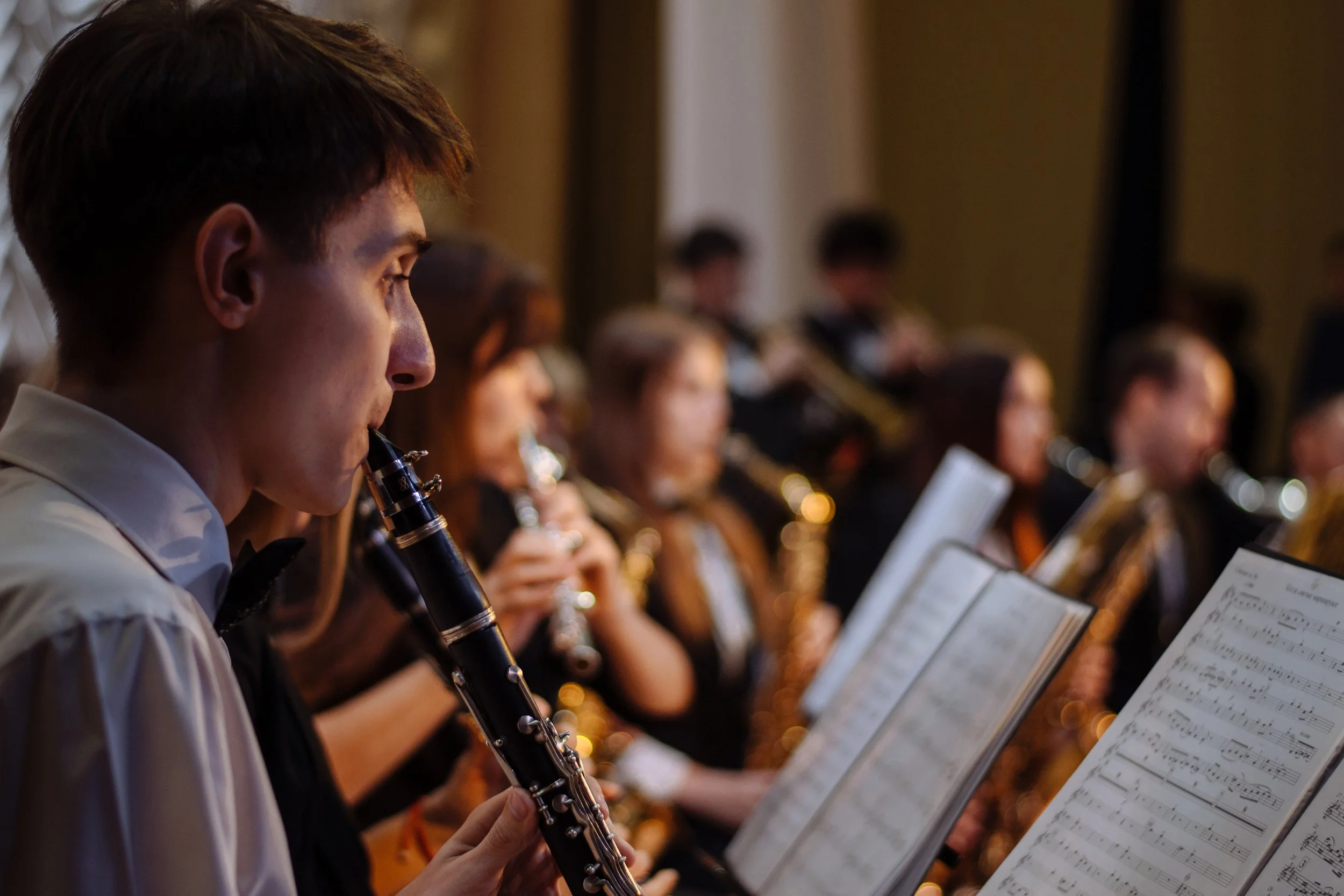 Young male musician playing the clarinet during a concert, with other musicians playing wind instruments in the background.