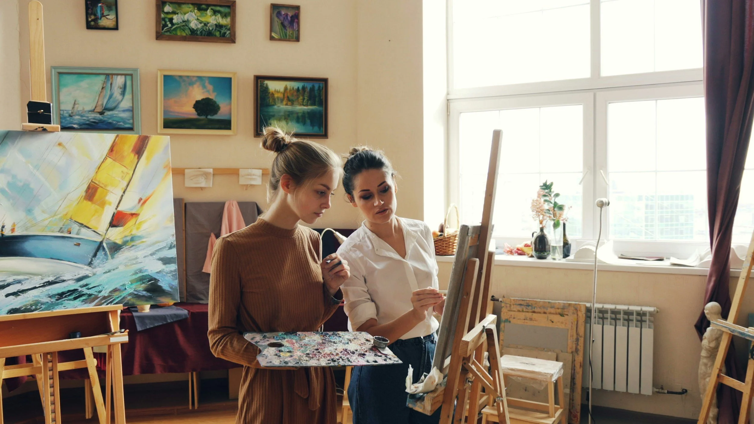 Two women are painting art in a well-lit art studio with paintings and windows in the background.