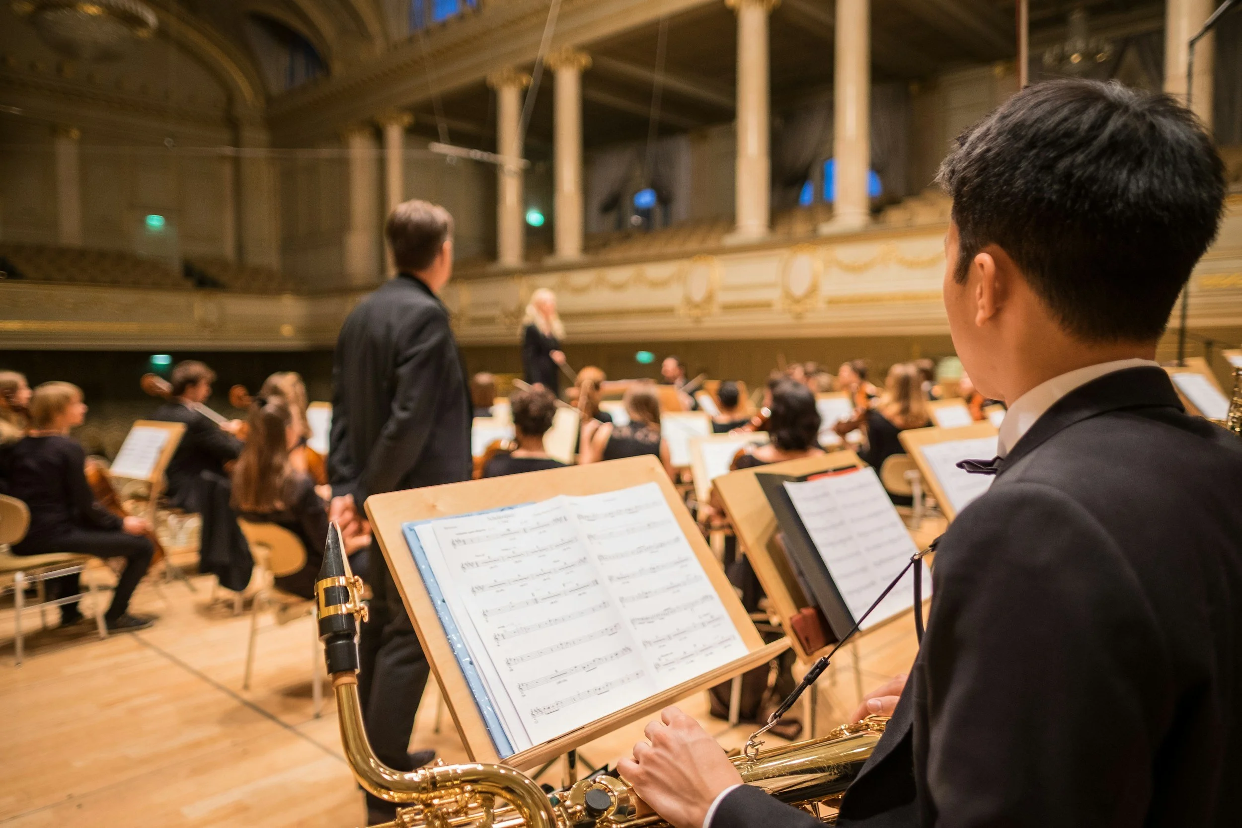 A performer playing a saxophone during an orchestral concert in a grand concert hall, with musicians and an conductor in the background.
