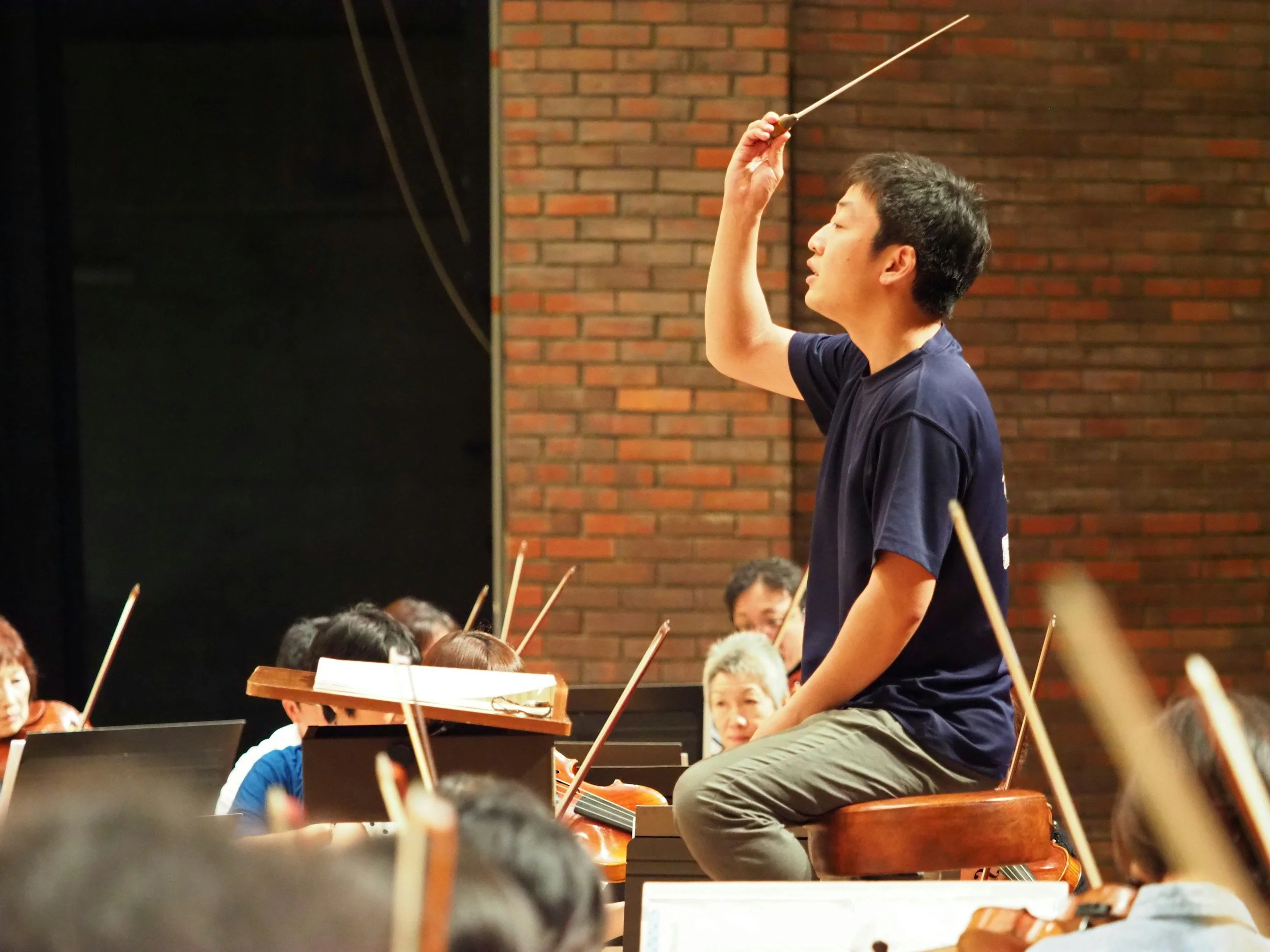 A young man with dark hair, wearing a navy blue T-shirt and khaki pants, conducts an orchestra during a performance inside a venue with brick walls. He is using a baton and is seated on a wooden chair. Several musicians with music stands and sheet music are visible around him.