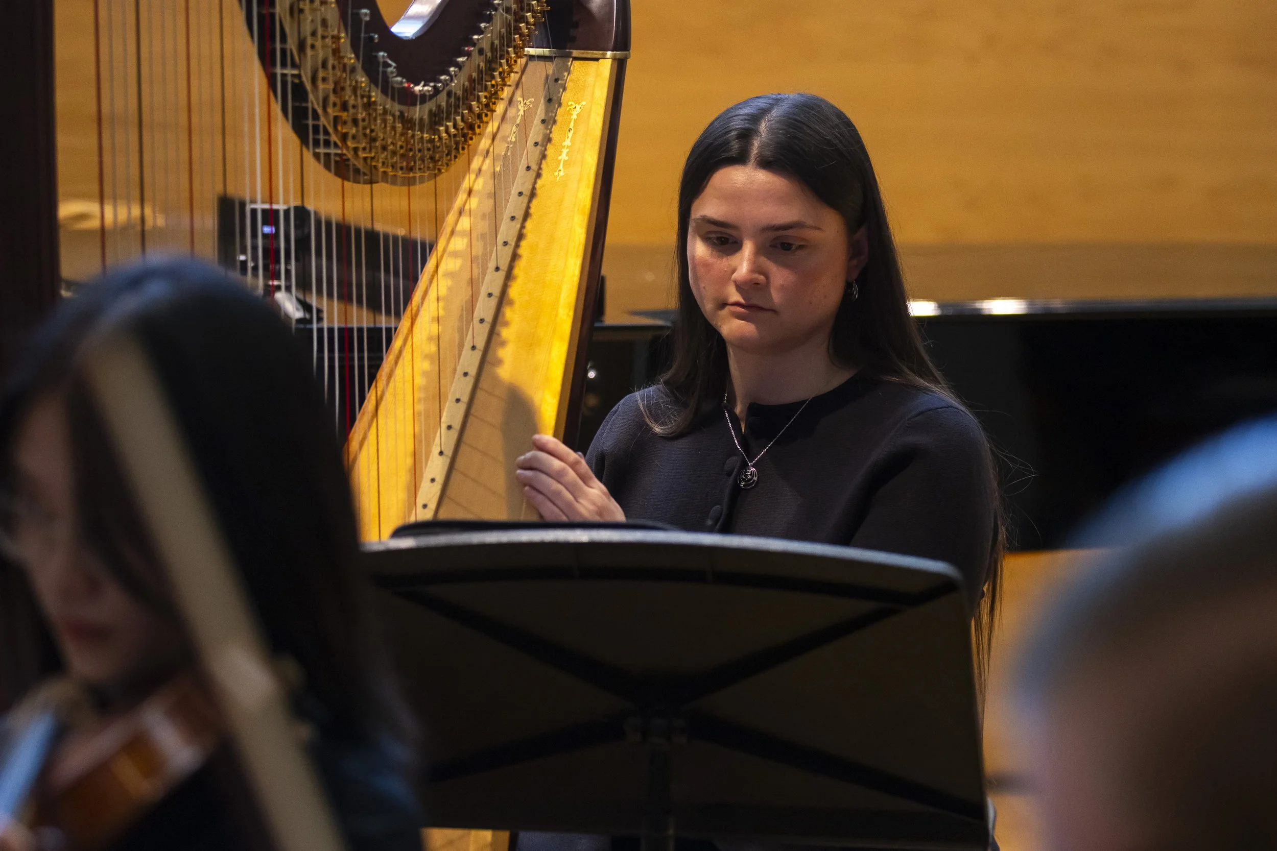 A young woman with long dark hair playing a harp in a performance or rehearsal space with a warm yellow background.