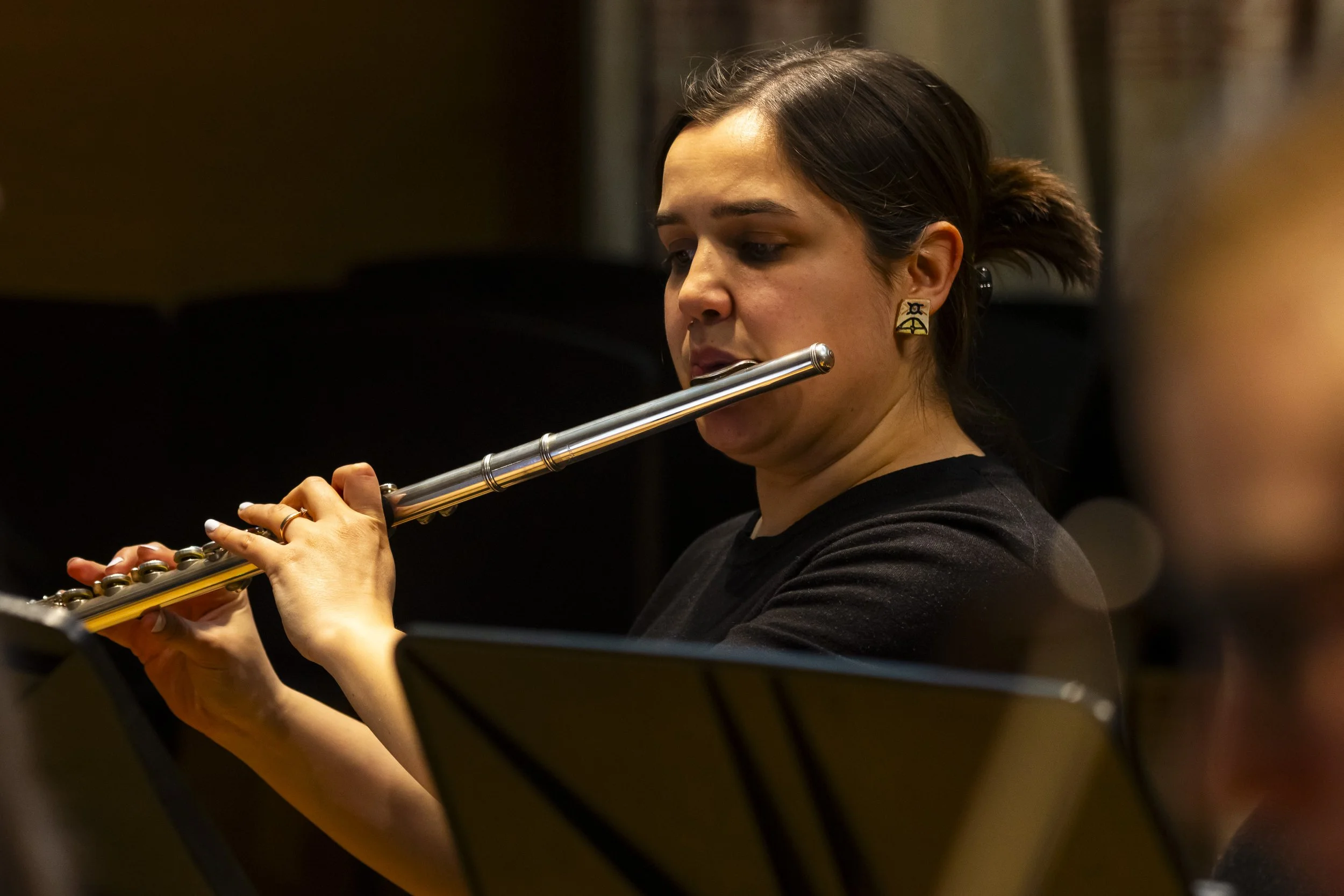A woman with dark hair playing a silver flute, wearing earrings, in a dark room or concert hall.