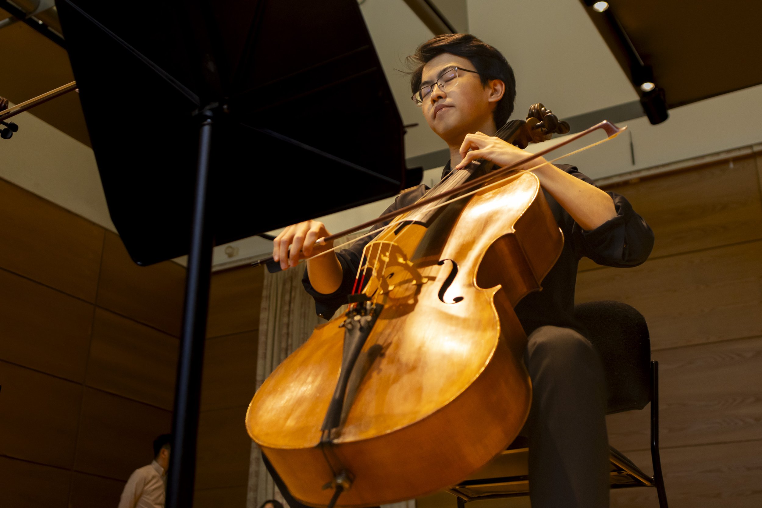 A man playing a cello with eyes closed, in a performance setting, with a music stand in the foreground and ceiling lights.
