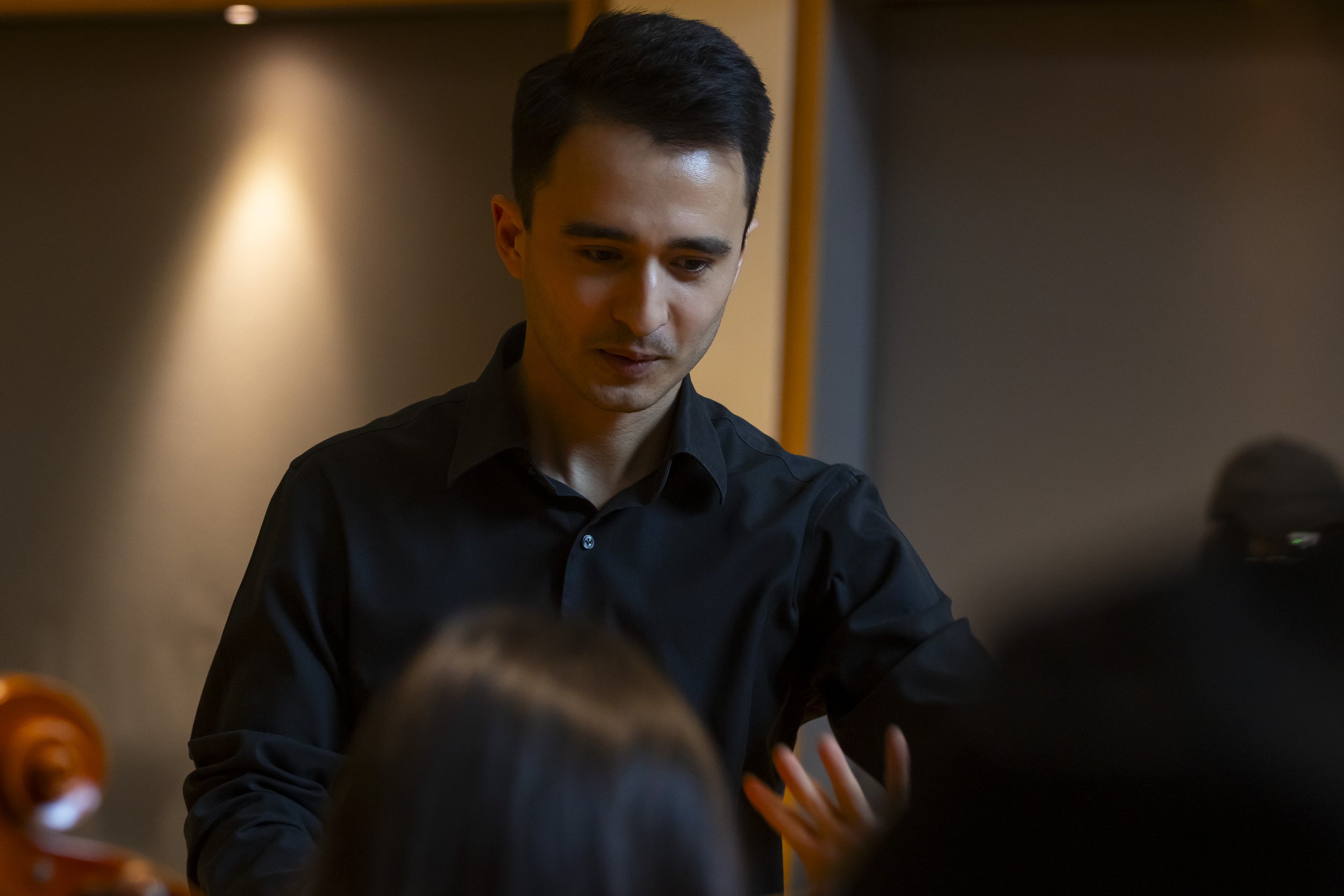 A young man with dark hair wearing a black shirt, looking down, in an indoor setting with warm lighting.