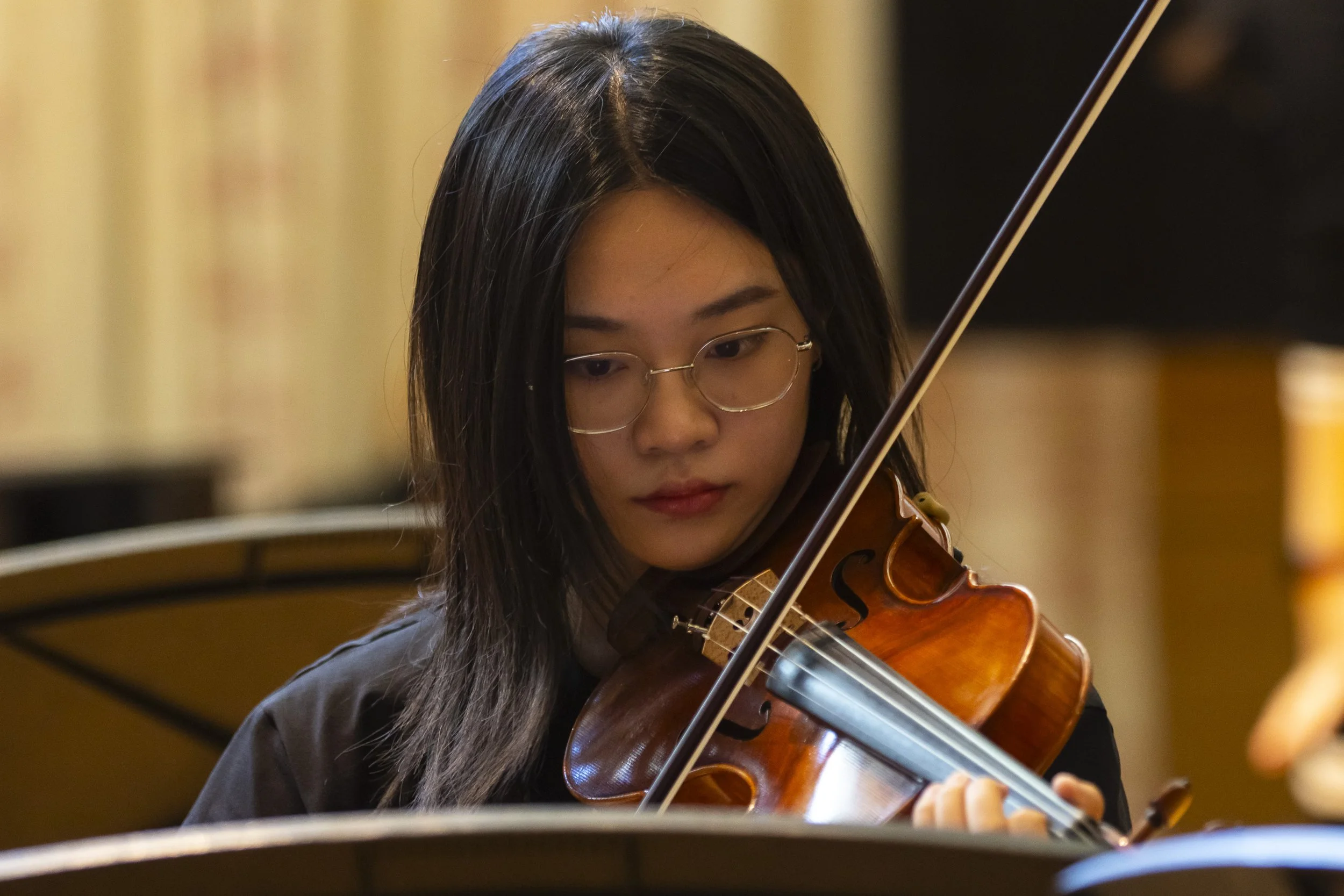 A young woman with long black hair and glasses playing a violin in a concert hall.