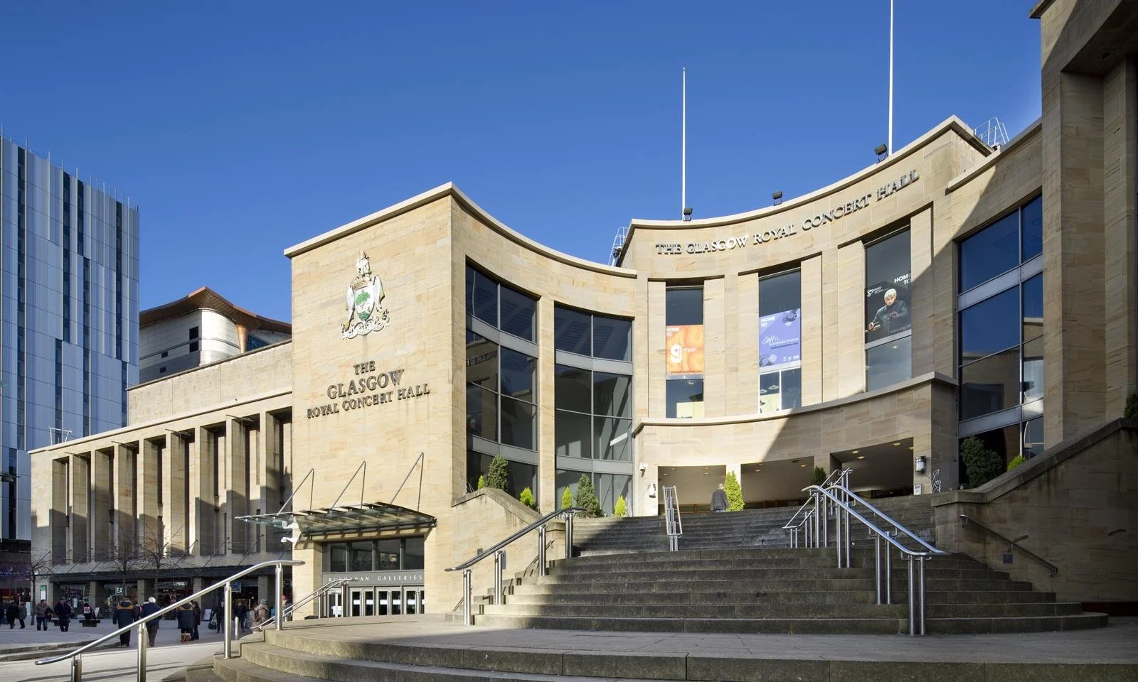 Exterior view of The Glasgow Royal Concert Hall, a modern beige building with large glass windows and steps leading up to the entrance, located in an urban area.