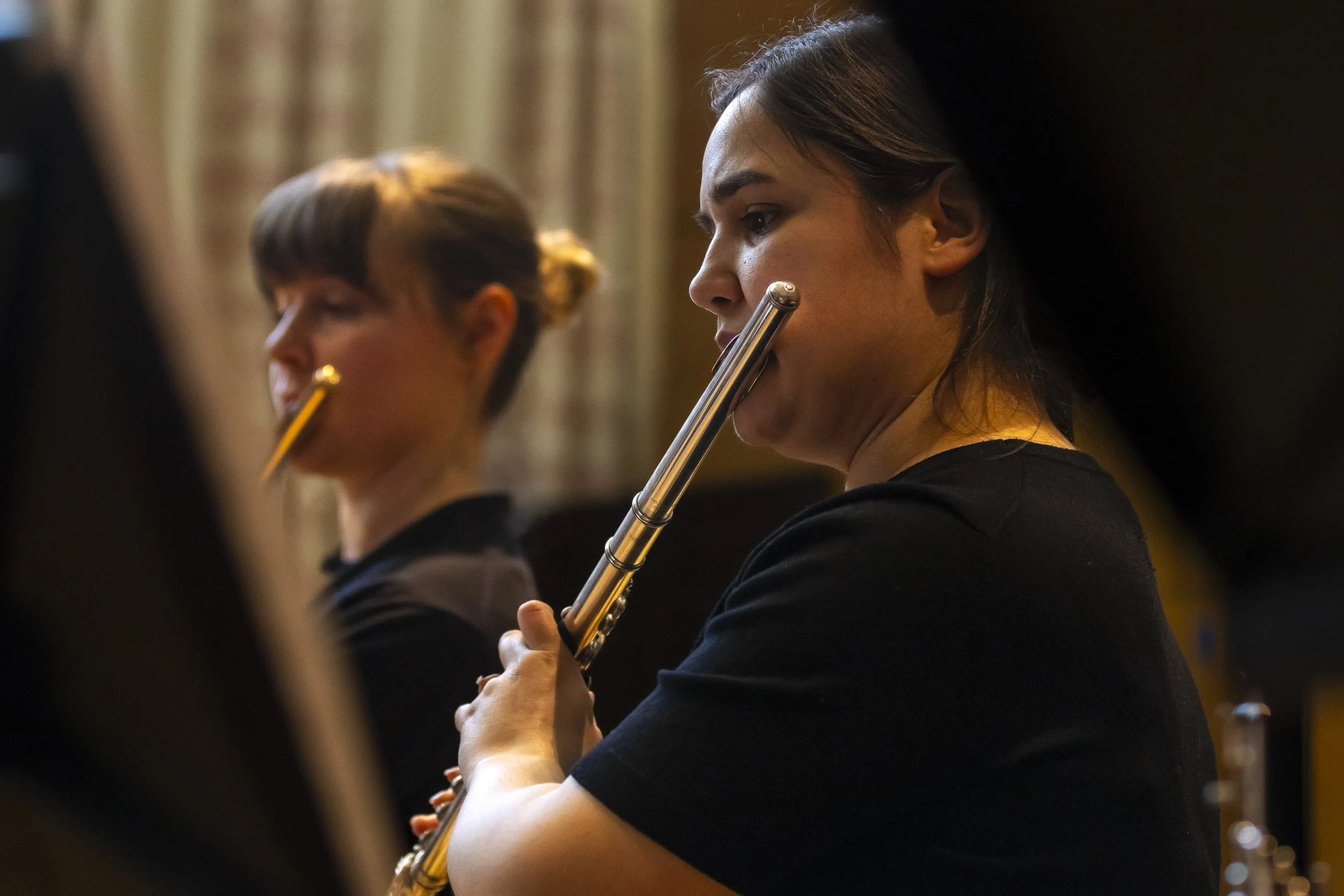 A woman playing the flute during a performance, with other musicians out of focus in the background.
