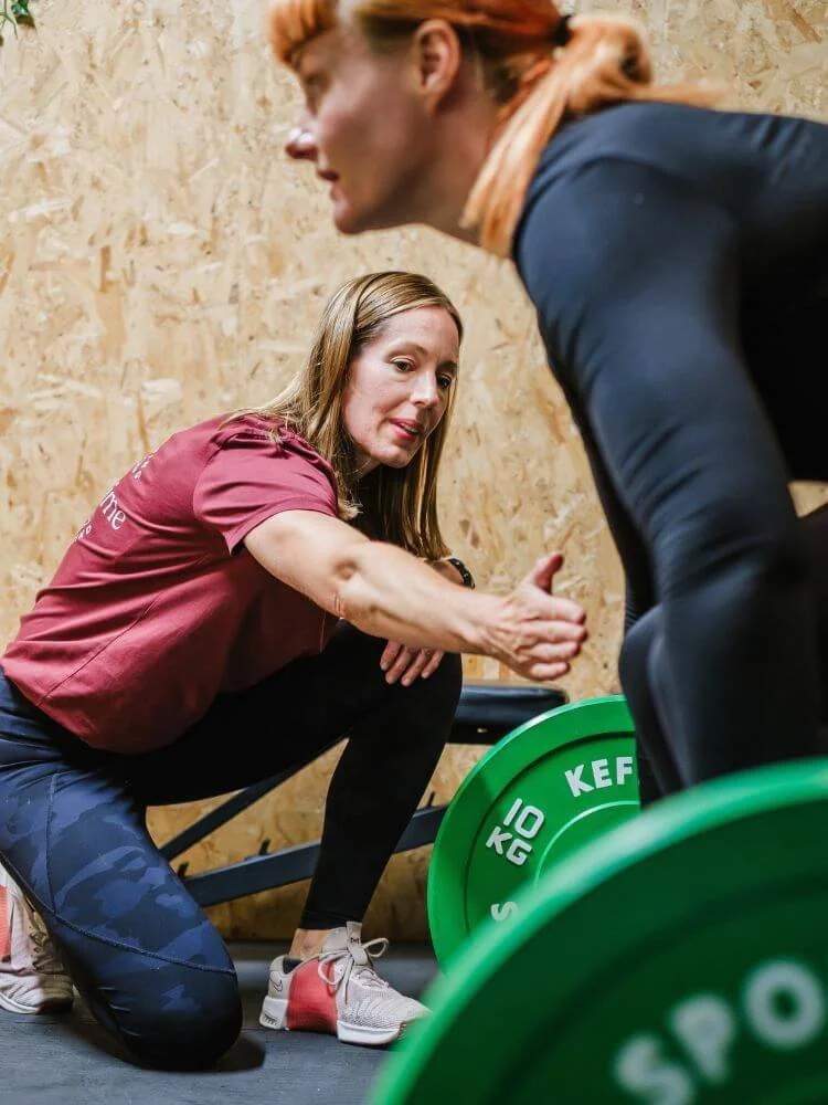A Personal Trainer in athletic wear kneeling and instructing another woman who is preparing to lift a barbell with green weights, inside a gym.