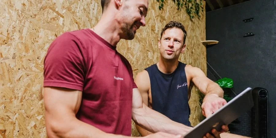 Two men in casual athletic clothing talking and looking at a tablet in a gym or workout space.