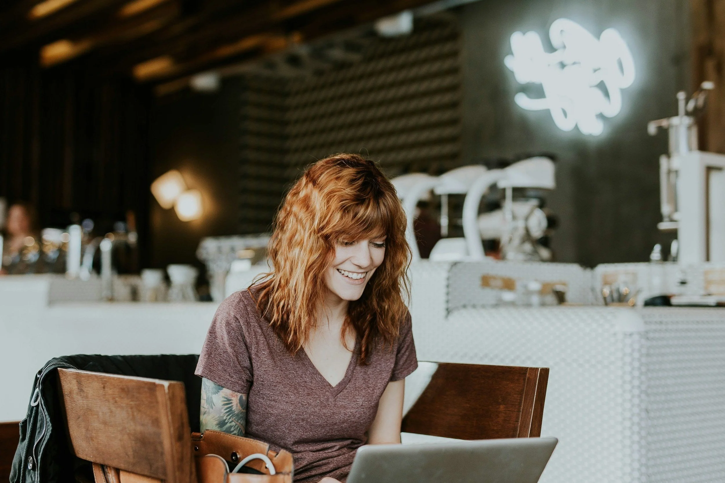 Mindset Coaching client with wavy hair smiling while using a laptop in a cafe.