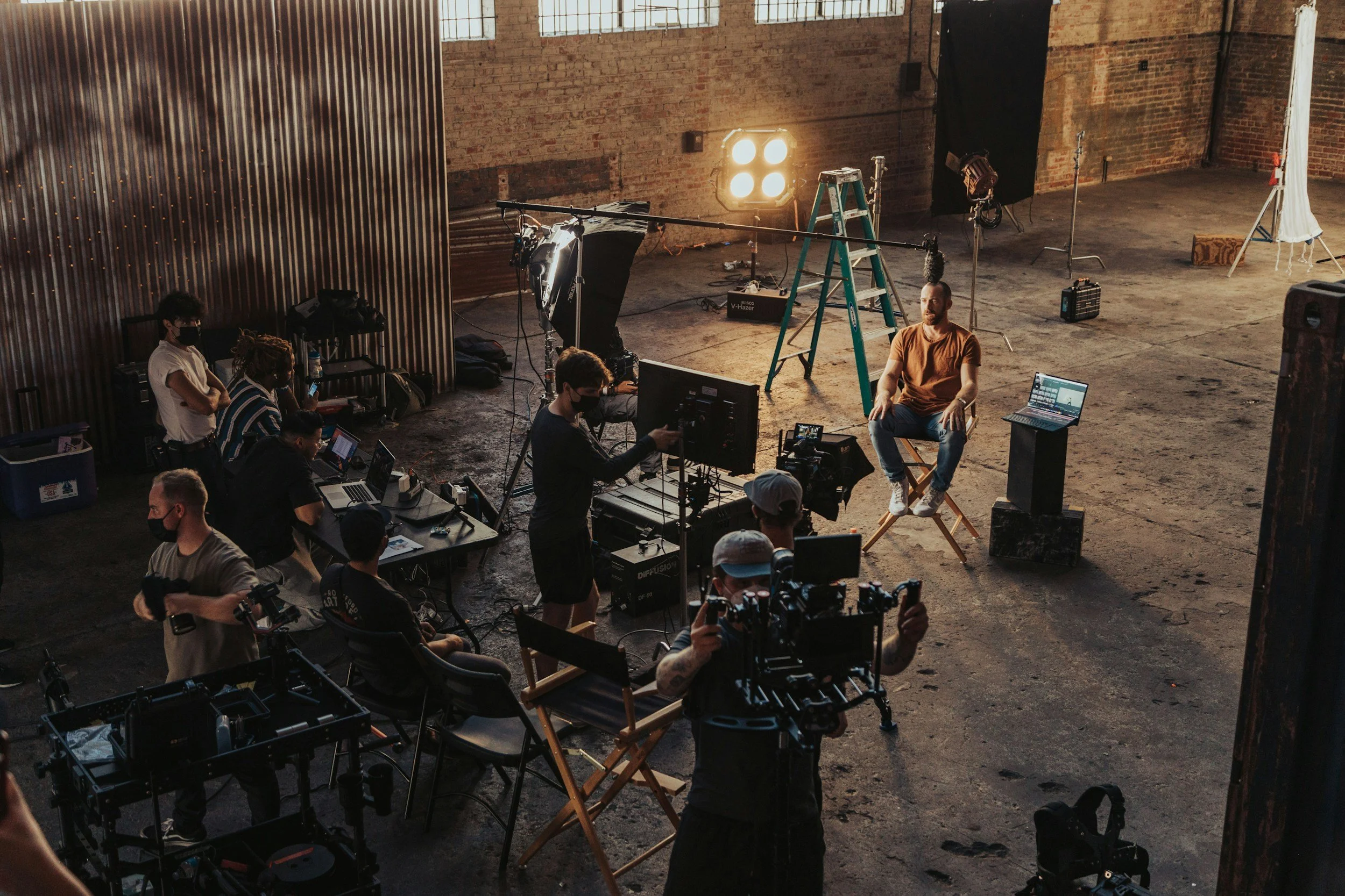 A behind-the-scenes view of a film or video shoot inside an industrial-style brick building with large windows, showing crew members and a man sitting on a director's chair being filmed.