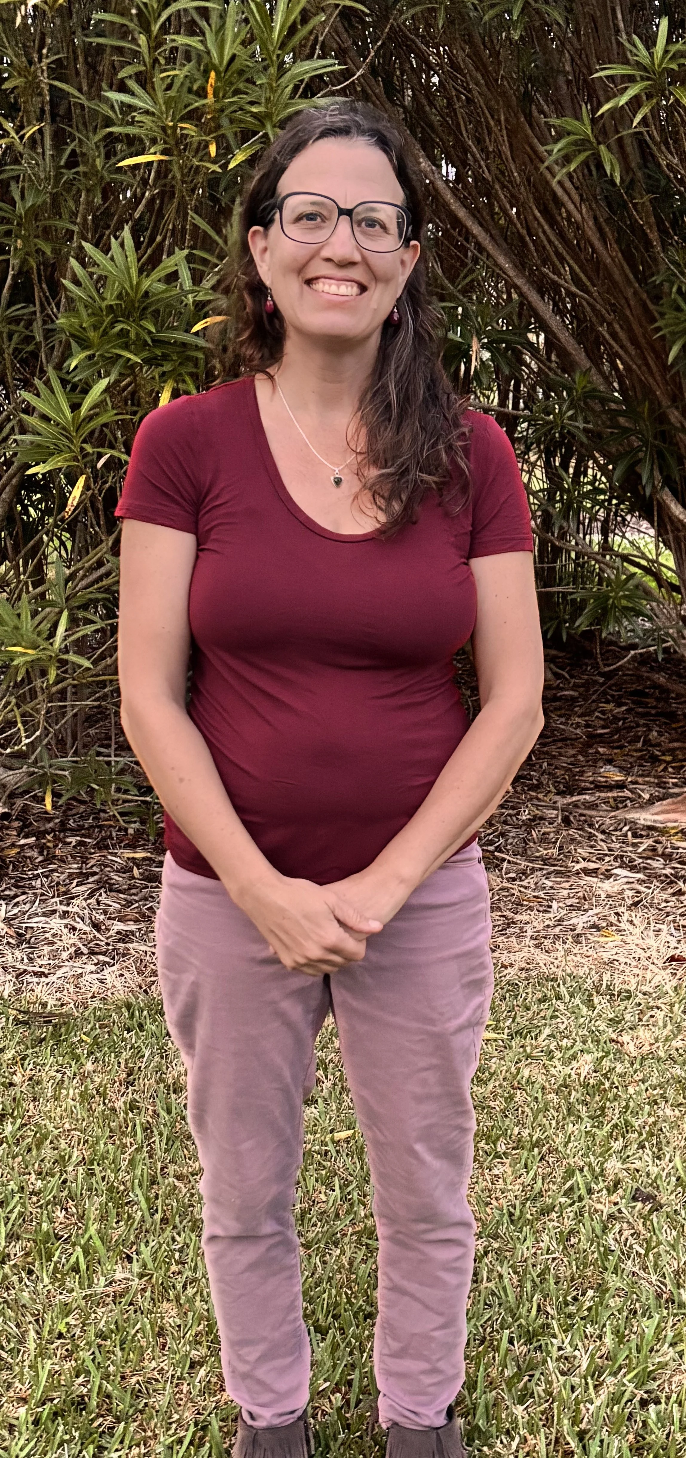 A woman with glasses, long brown hair, and earrings, smiling and standing outdoors in front of green shrubbery, wearing a maroon T-shirt and light pink pants.