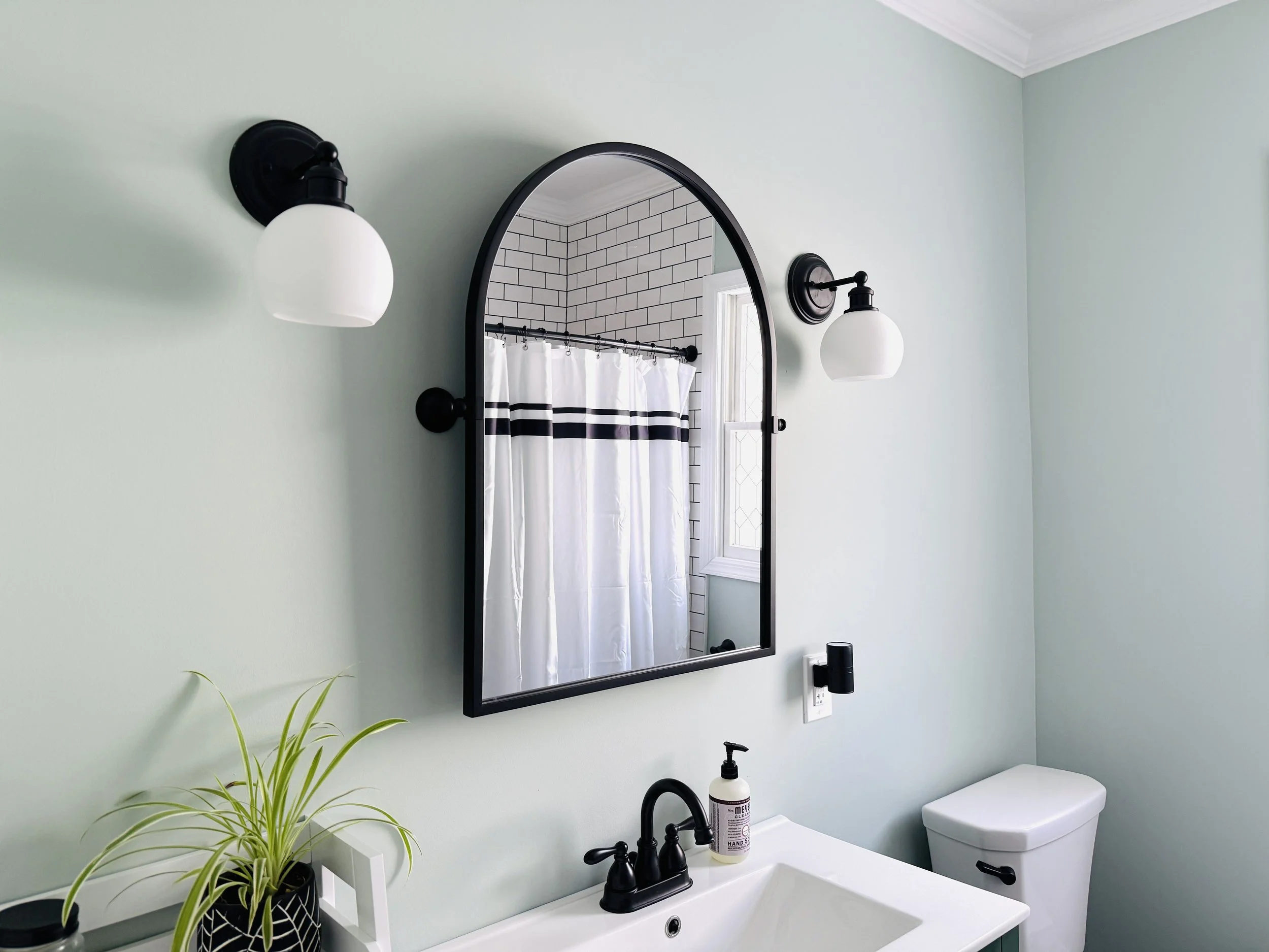 A bathroom with pale green walls, a black-framed mirror above a white sink, two black wall-mounted light fixtures with white globes on either side of the mirror, a potted plant with long green leaves, a soap dispenser, and a white toilet. A reflection in the mirror shows a shower with a white curtain and black stripes, and a window with white framing and frosted glass.