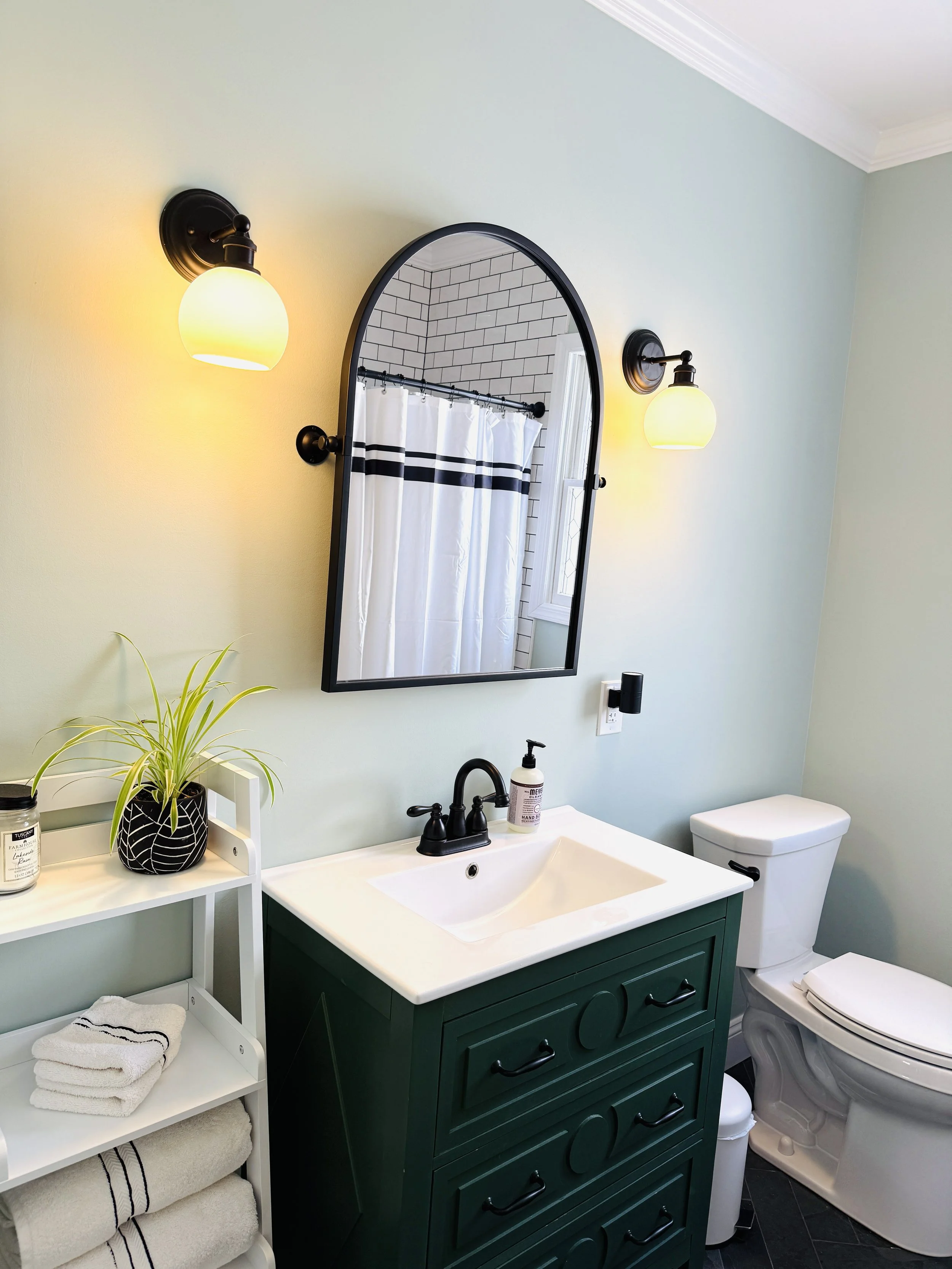 A modern bathroom with a green vanity, white countertop, and black hardware. A mirror above the sink, two wall-mounted lights, a white shelf with towels and a plant, and a white toilet are visible.