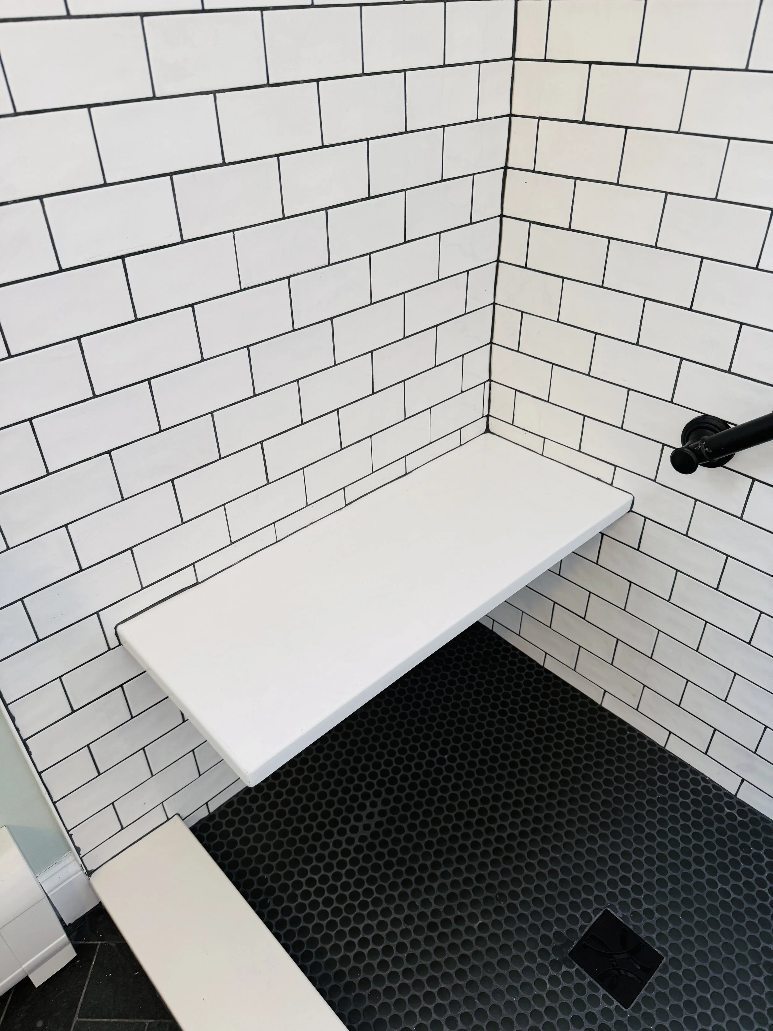 Shower with white subway tile walls, white bench, black circular drain, black shower handle, and black hexagonal floor tiles.
