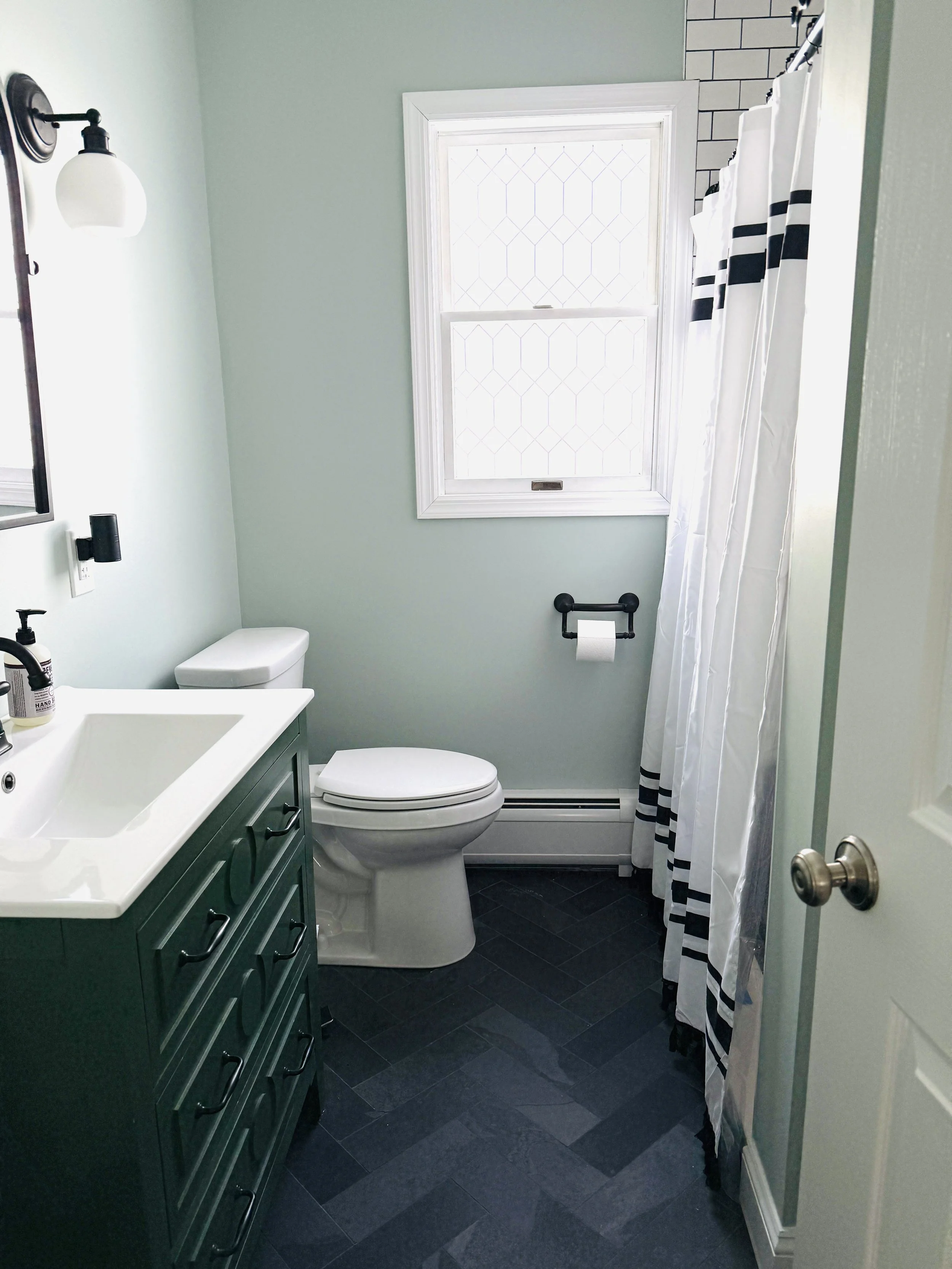 Small bathroom with a dark tiled floor, white toilet, teal vanity with a sink, wall-mounted black faucet and light fixture, white window with patterned glass, and a shower with a black and white striped curtain.
