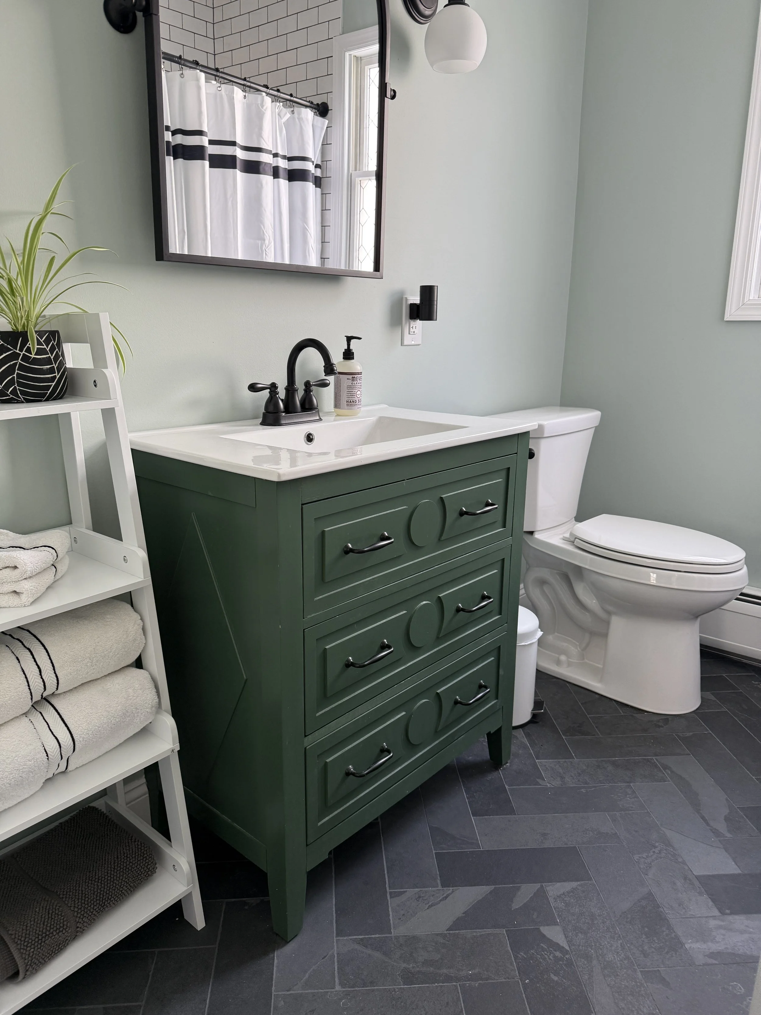 Bathroom with green vanity, white sink, black faucet, mirror, white toilet, black and white shower curtain, and black and white towels on a white shelf, with dark gray tiled floor.