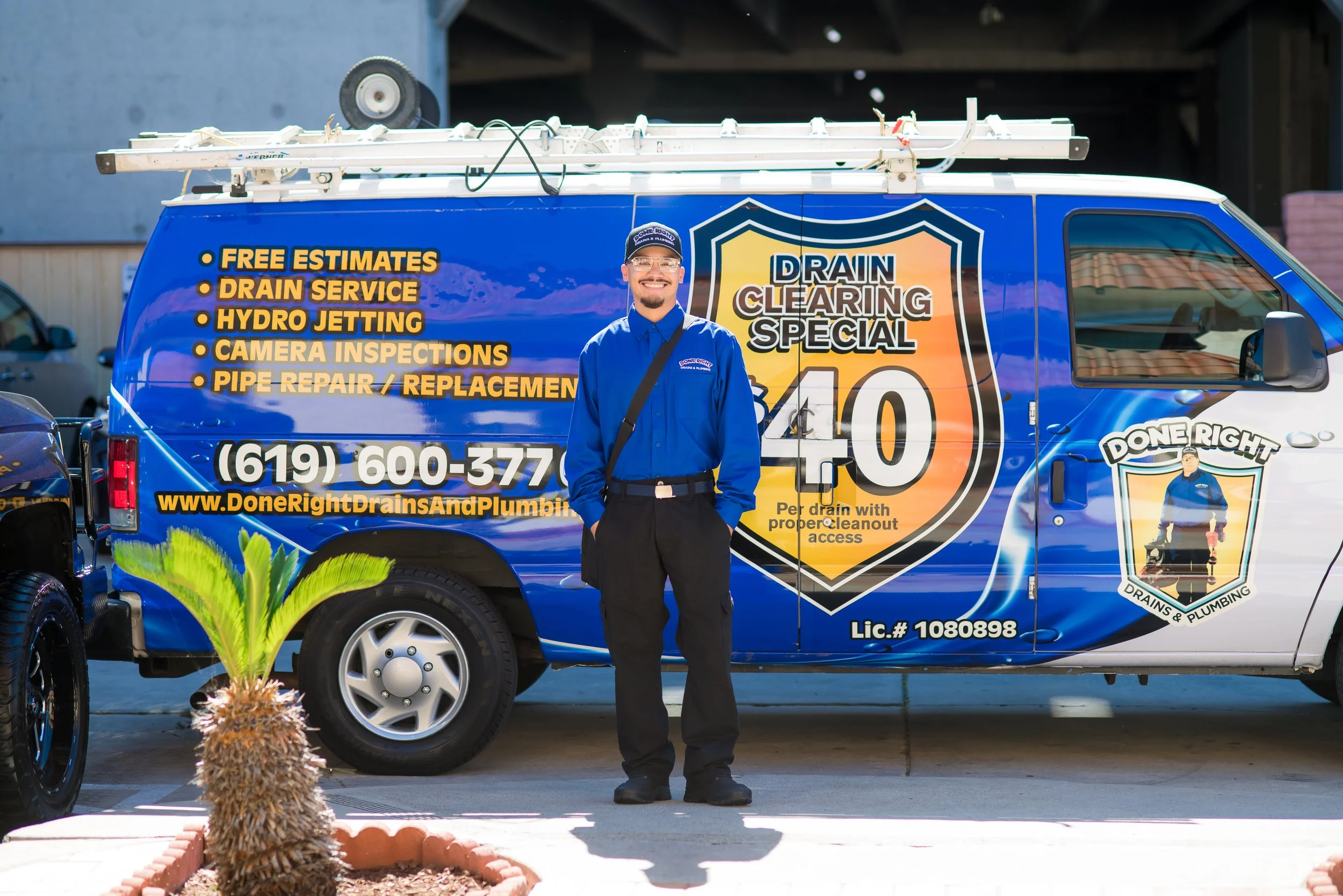 Smiling man in blue uniform standing in front of a blue drain cleaning service van. The van has a logo and list of services, including free estimates, drain service, hydro jetting, camera inspections, and pipe repair. The background shows a building and a parking lot.