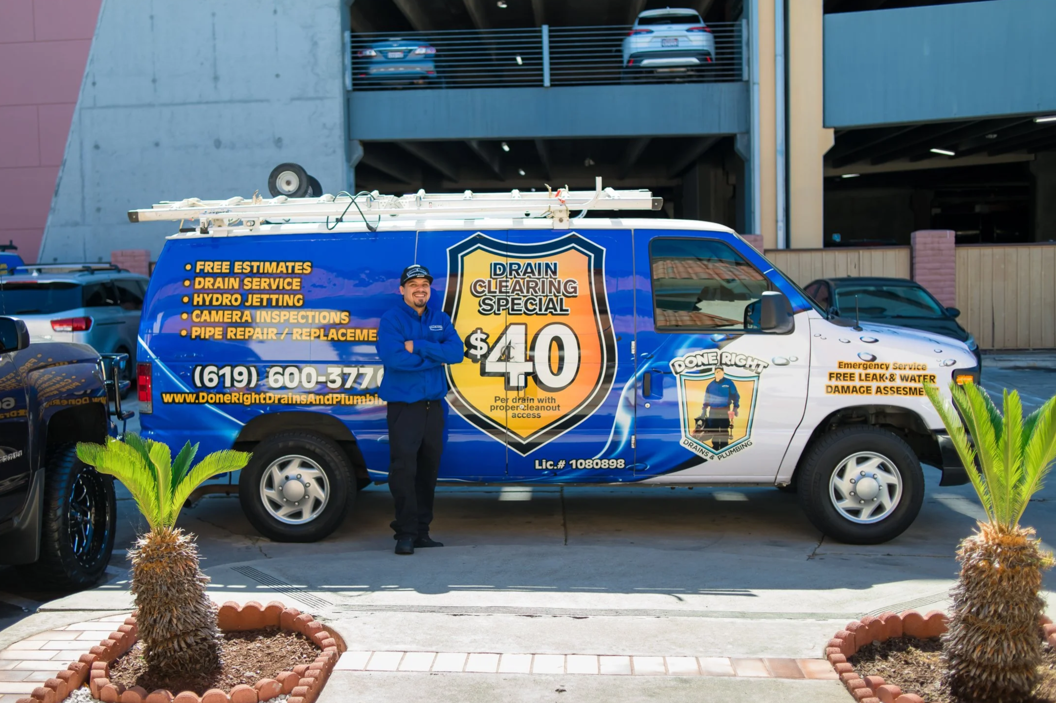 A man standing in front of a blue service van advertising drain services, parked in a parking lot with decorative plants and trees in the foreground.
