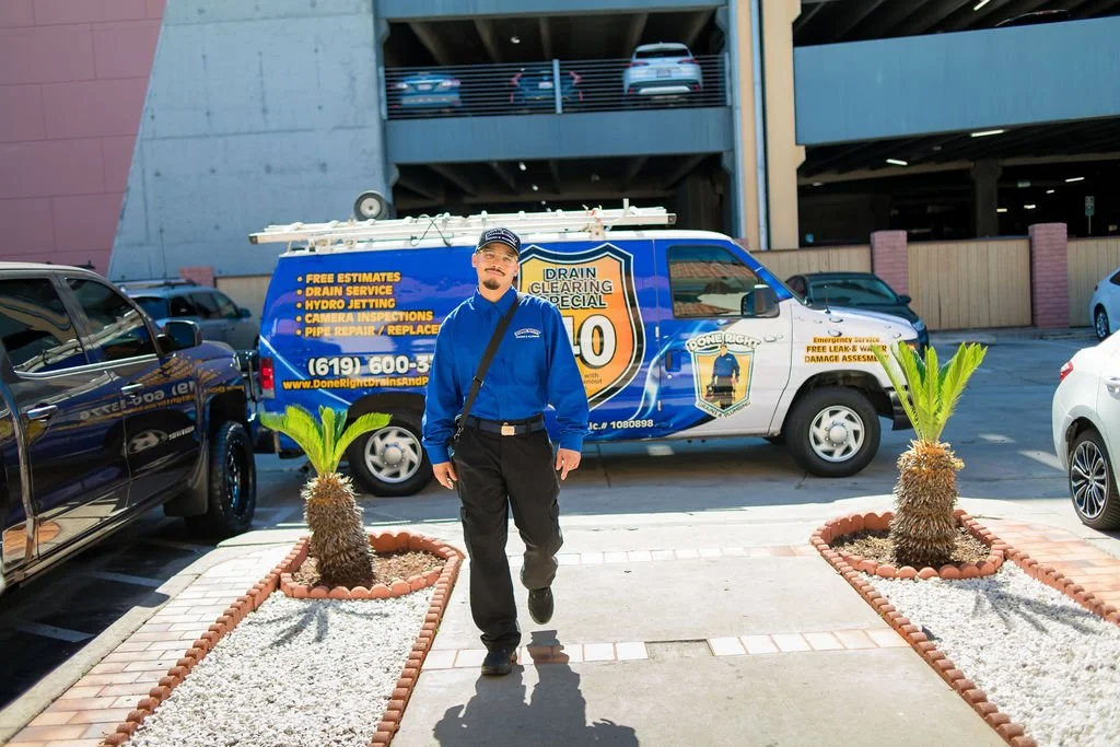 A worker in blue uniform standing in front of a plumbing and drain cleaning service vehicle in an outdoor parking lot.