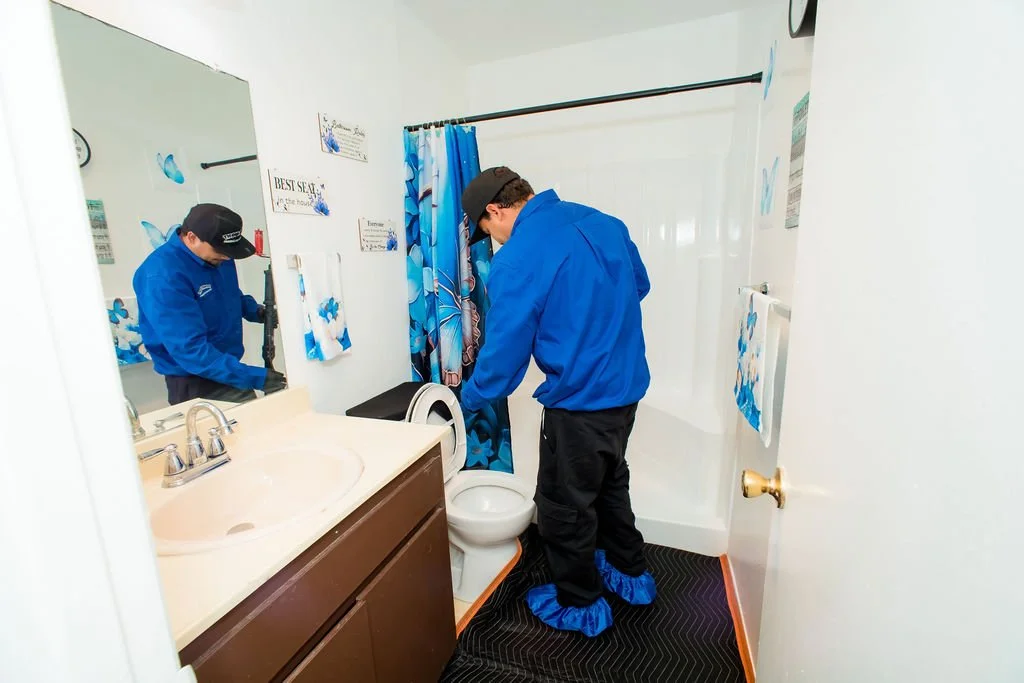 A man wearing a black cap, blue jacket, black pants, and blue shoe covers cleaning a toilet in a small bathroom with a shower curtain, towel, and mirror.