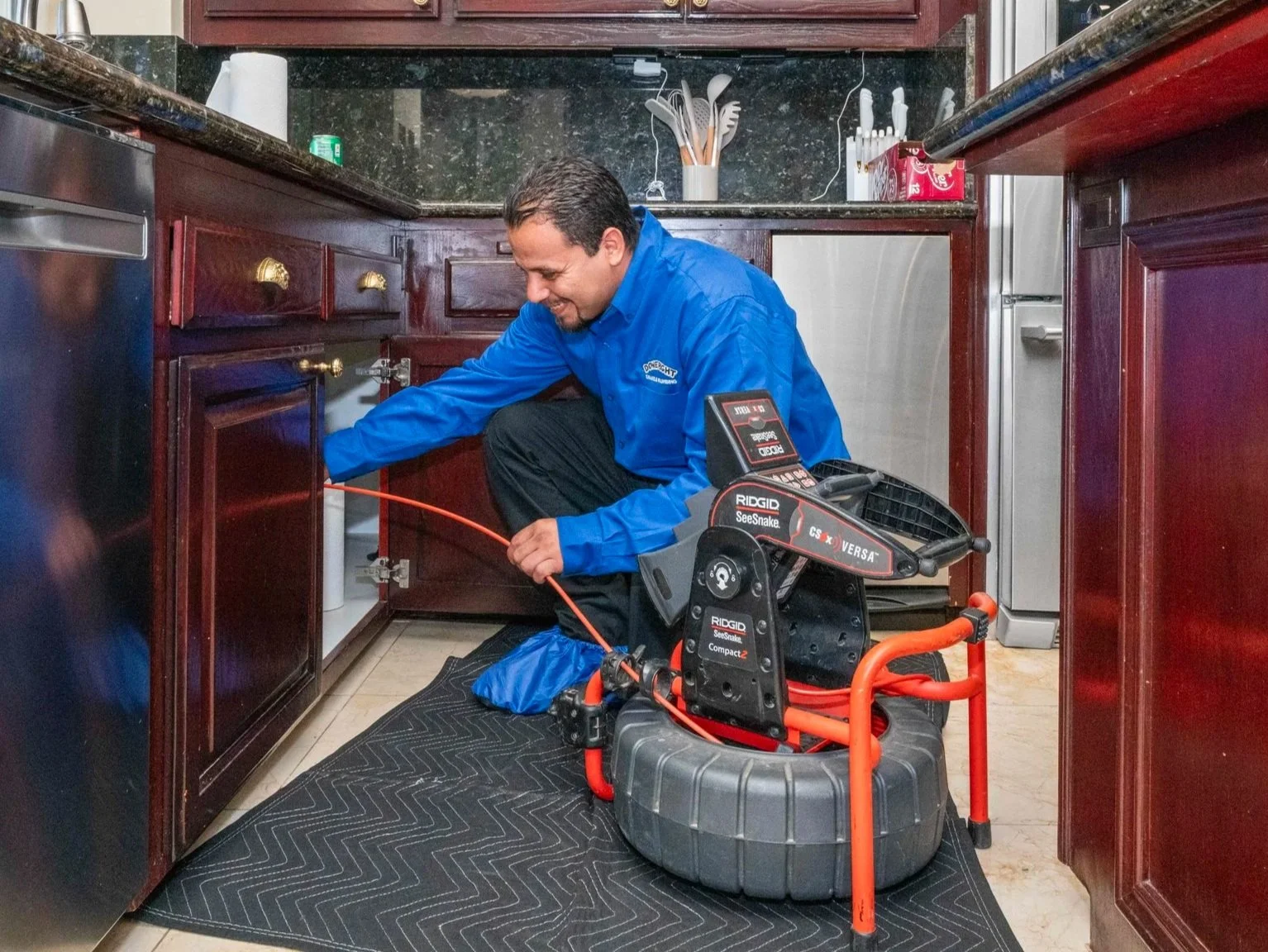 A technician in a blue uniform inspecting or repairing a kitchen cabinet using specialized equipment. The technician is kneeling on a black protective mat with tools and a mechanical device around them.