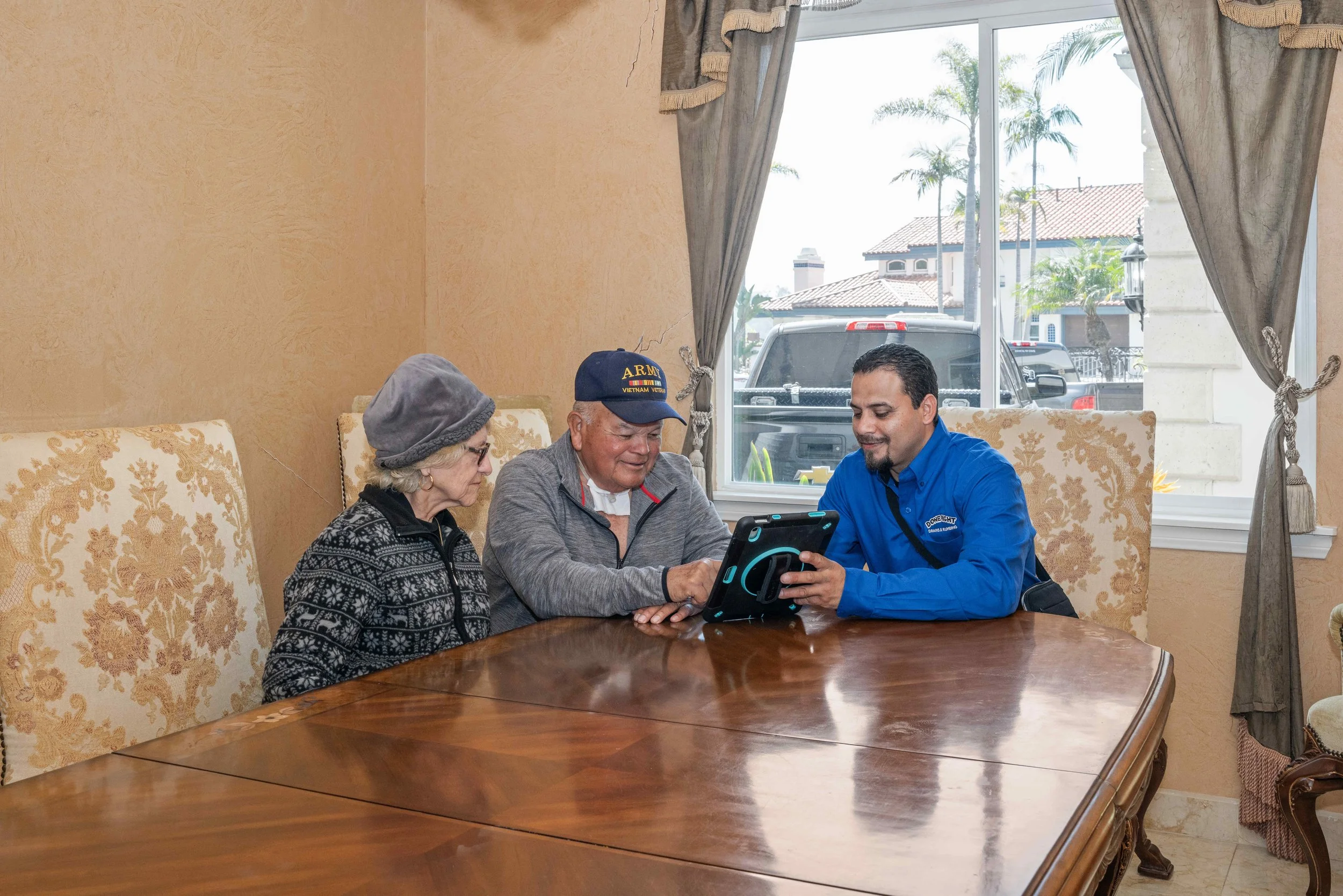 Three people sitting around a wooden table, looking at a device together inside a room with large window and beige walls.