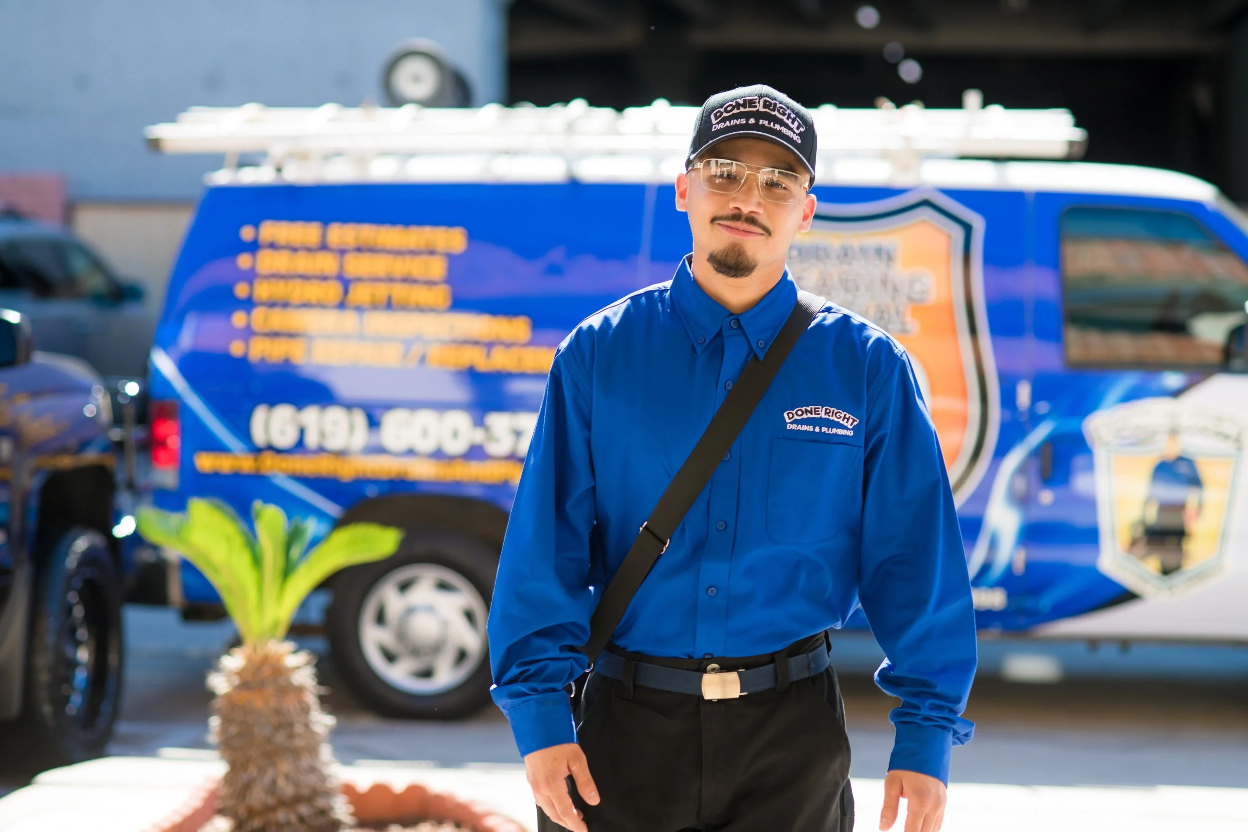 A man in a blue uniform with a black cap and safety glasses stands in front of a blue service vehicle with yellow and orange text. The vehicle advertises plumbing and drain services. The man has a goatee and a slight smile and is holding a smartphone in his right hand.